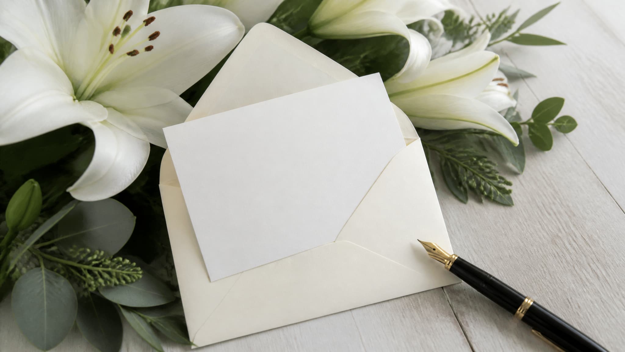 A small blank white florist card in a cream envelope resting on a spray of white lilies and greenery, a fountain pen lying beside it on a wooden surface