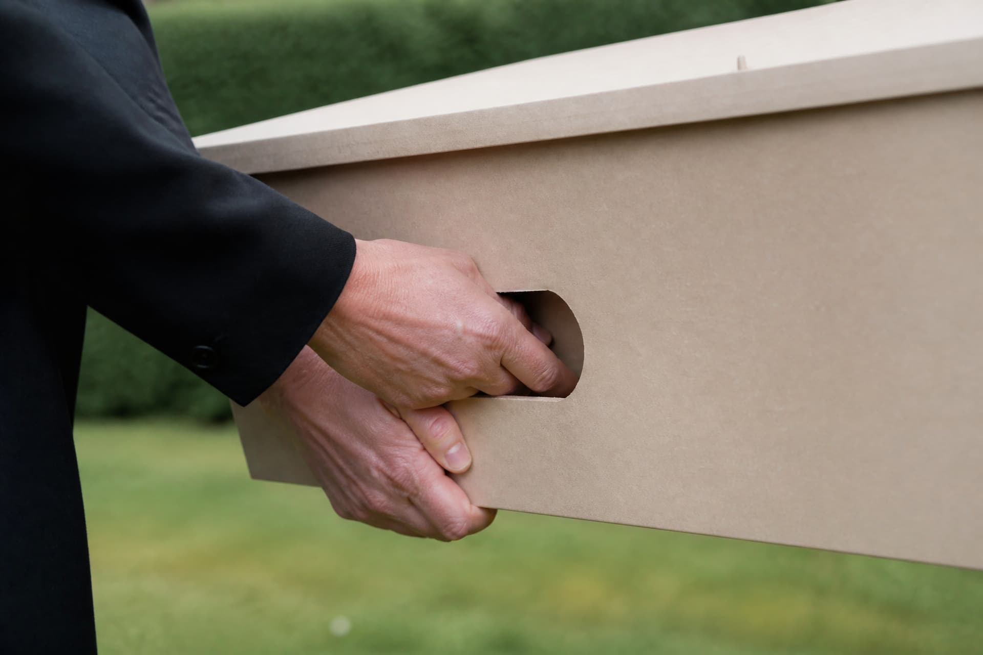 Close view of a cut-out oval handhold in the side of a pale cardboard coffin, two adult hands gripping it, one from above and one supporting underneath, dark sleeves entering from the left with grass out of focus behind