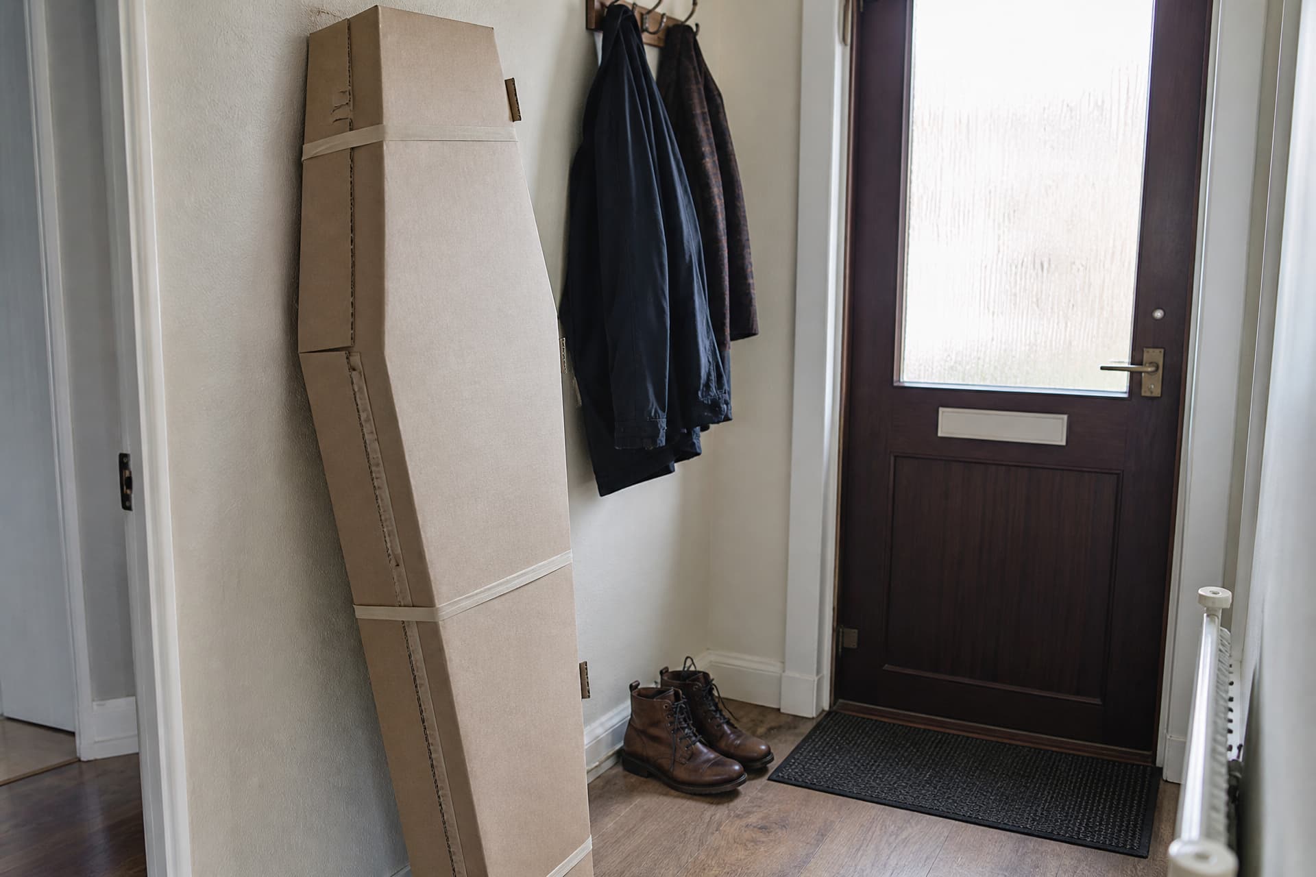 A plain tapered cardboard coffin leaning upright and still strapped against the wall of a domestic hallway, beside a front door with coats on a rack and a pair of boots on the floor