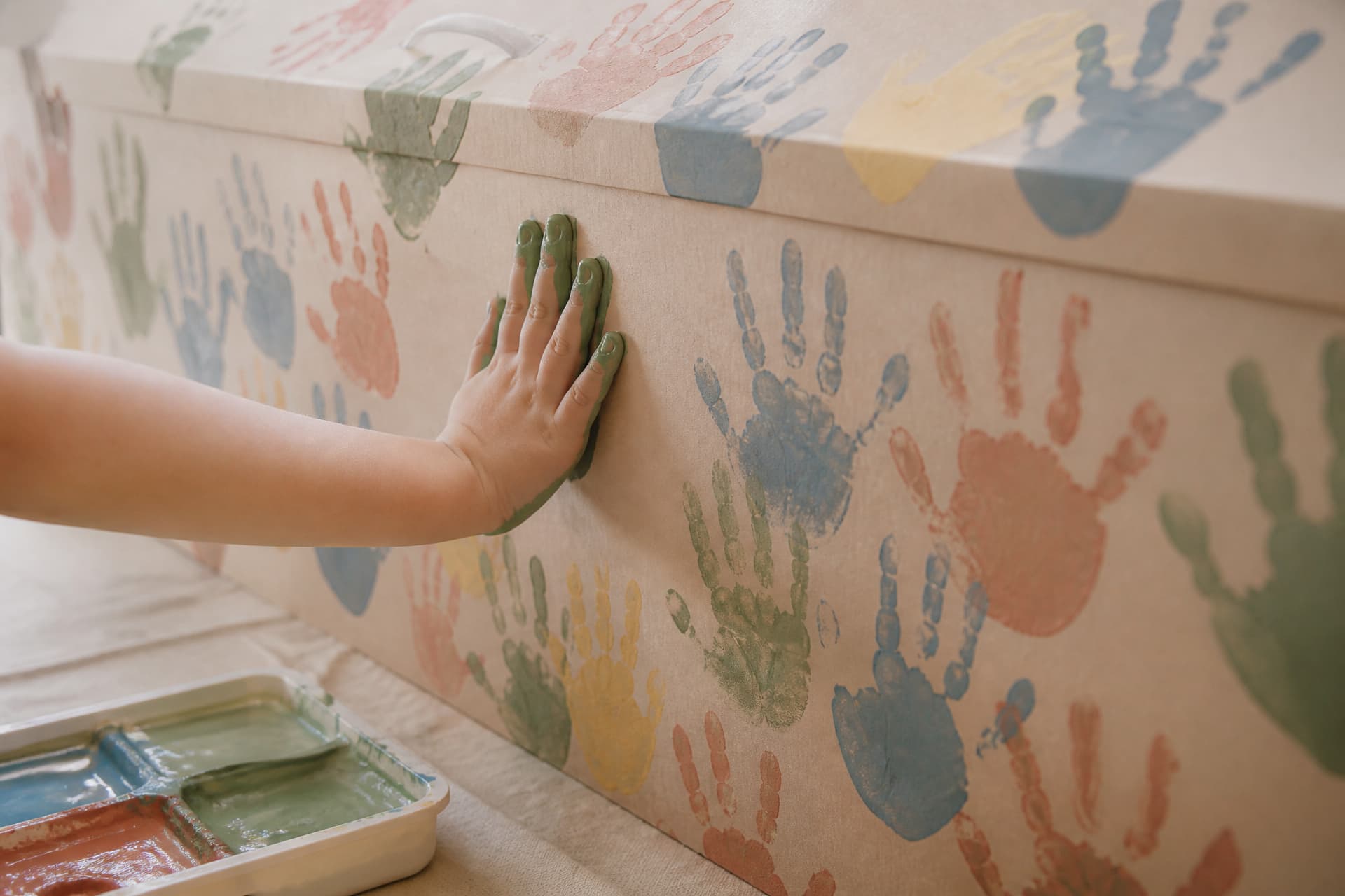 The side of a pale cardboard coffin covered in overlapping painted handprints in blue, green, yellow and terracotta, with a small child's hand pressing a freshly painted green palm onto a bare patch of board
