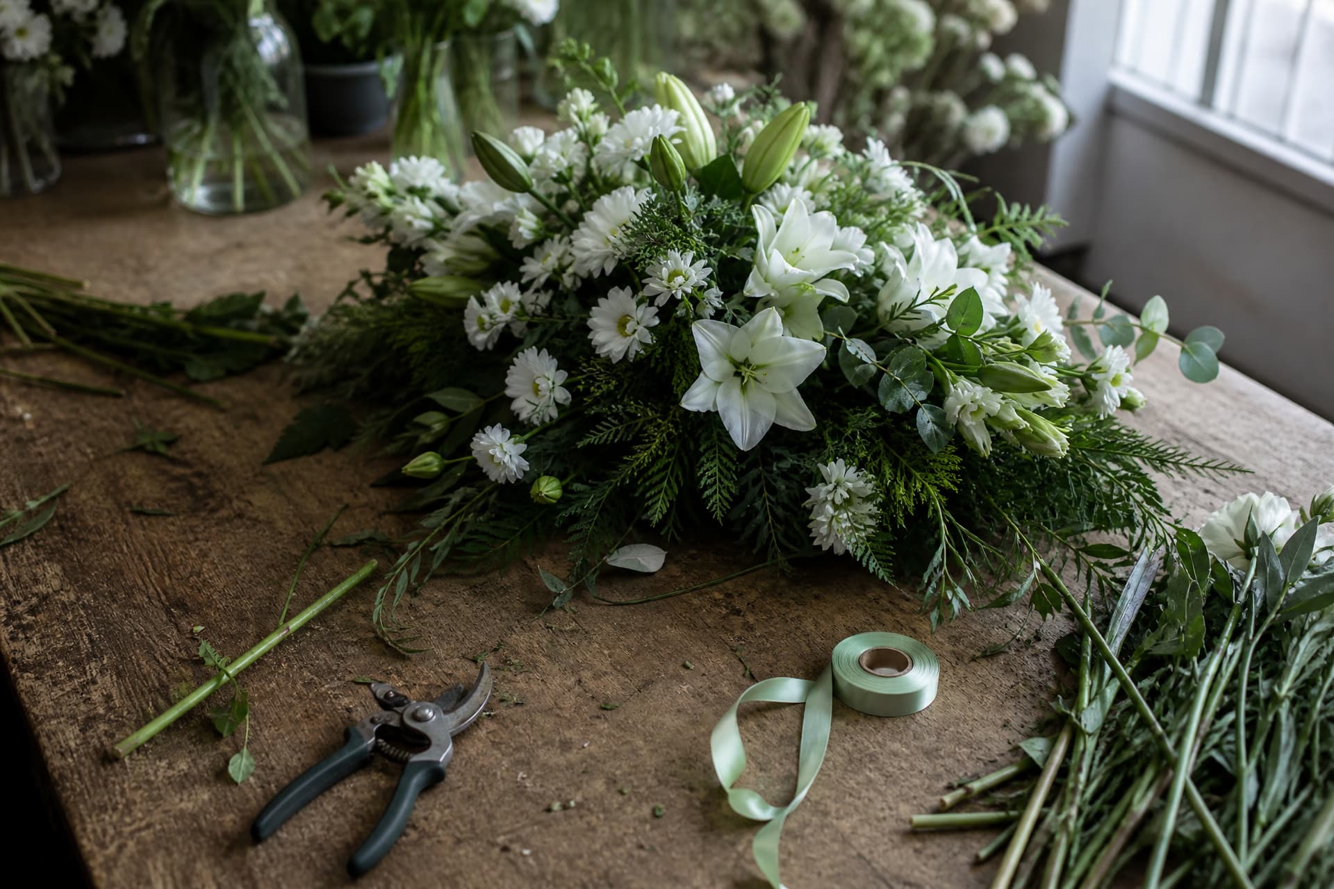 A florist's workbench seen from above, a funeral spray part-assembled in white and green, secateurs, a spool of ribbon and cut stems scattered across the wood