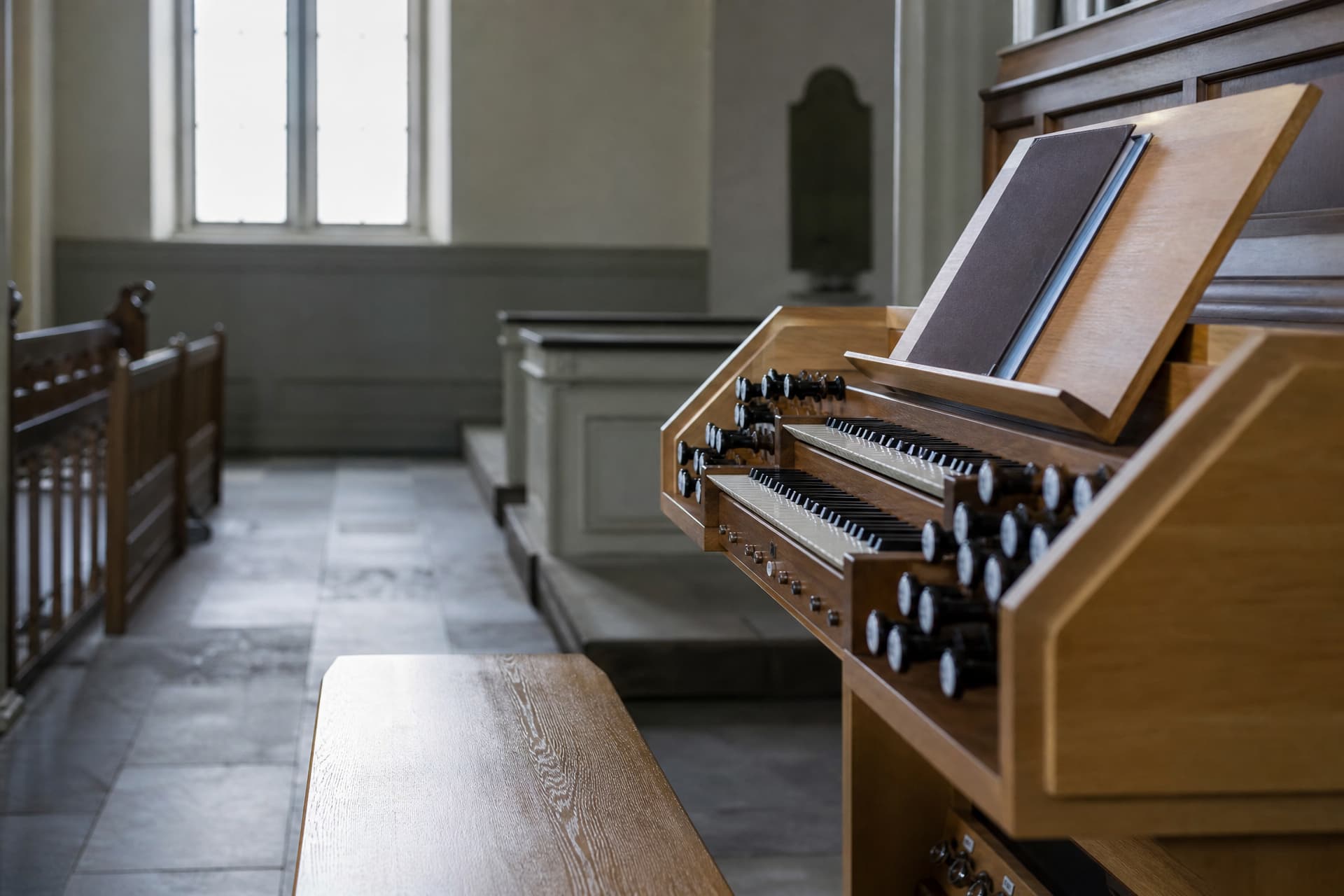 A pipe organ console in an empty parish church seen from the side, the bench empty and pushed back, sheet music closed on the stand, cool daylight through a plain window behind