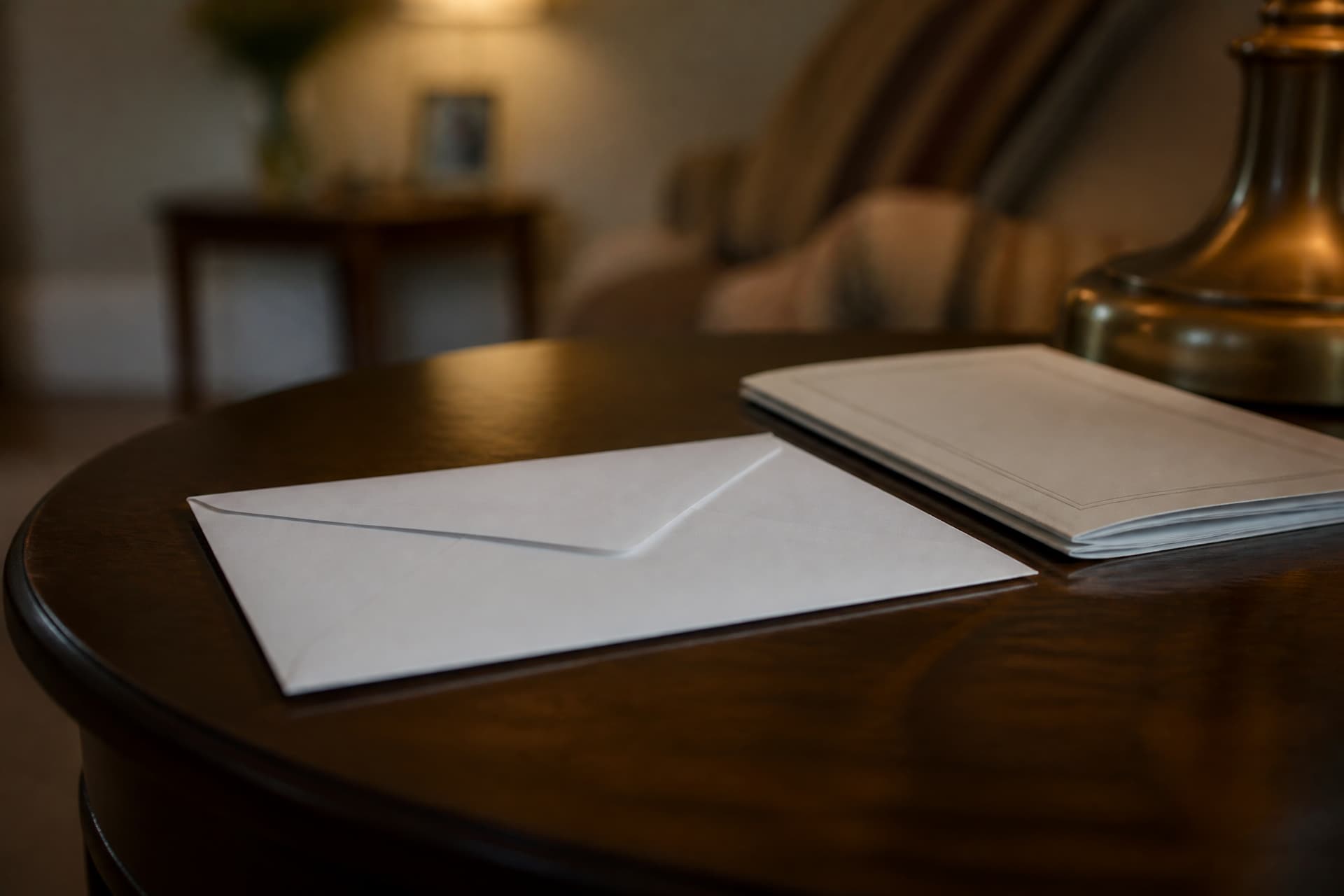A plain white envelope resting on a dark wooden side table beside a folded order of service, lit by a warm lamp, unmarked and sealed
