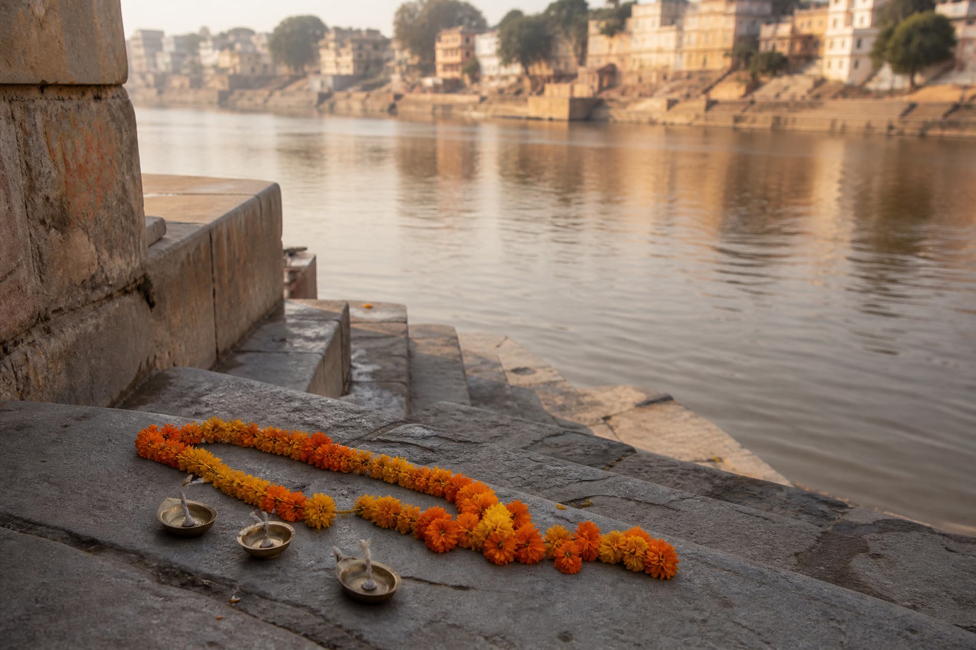 Worn stone steps of a riverside ghat leading down to the water in soft afternoon light, with marigold garlands and small brass lamps resting on the steps