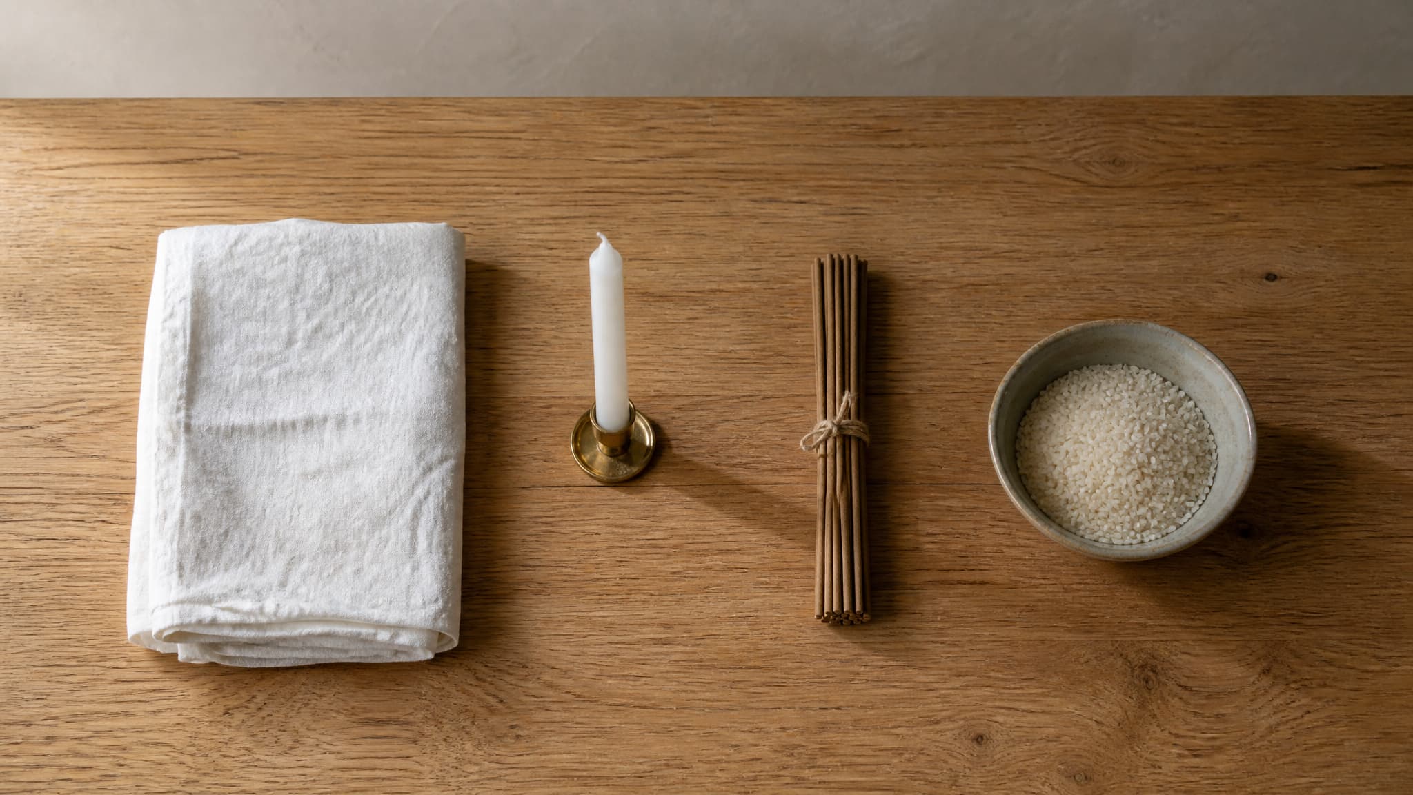 A plain oak table holding folded white linen, an unlit candle in a brass holder, a bundle of incense sticks and a shallow ceramic bowl of rice