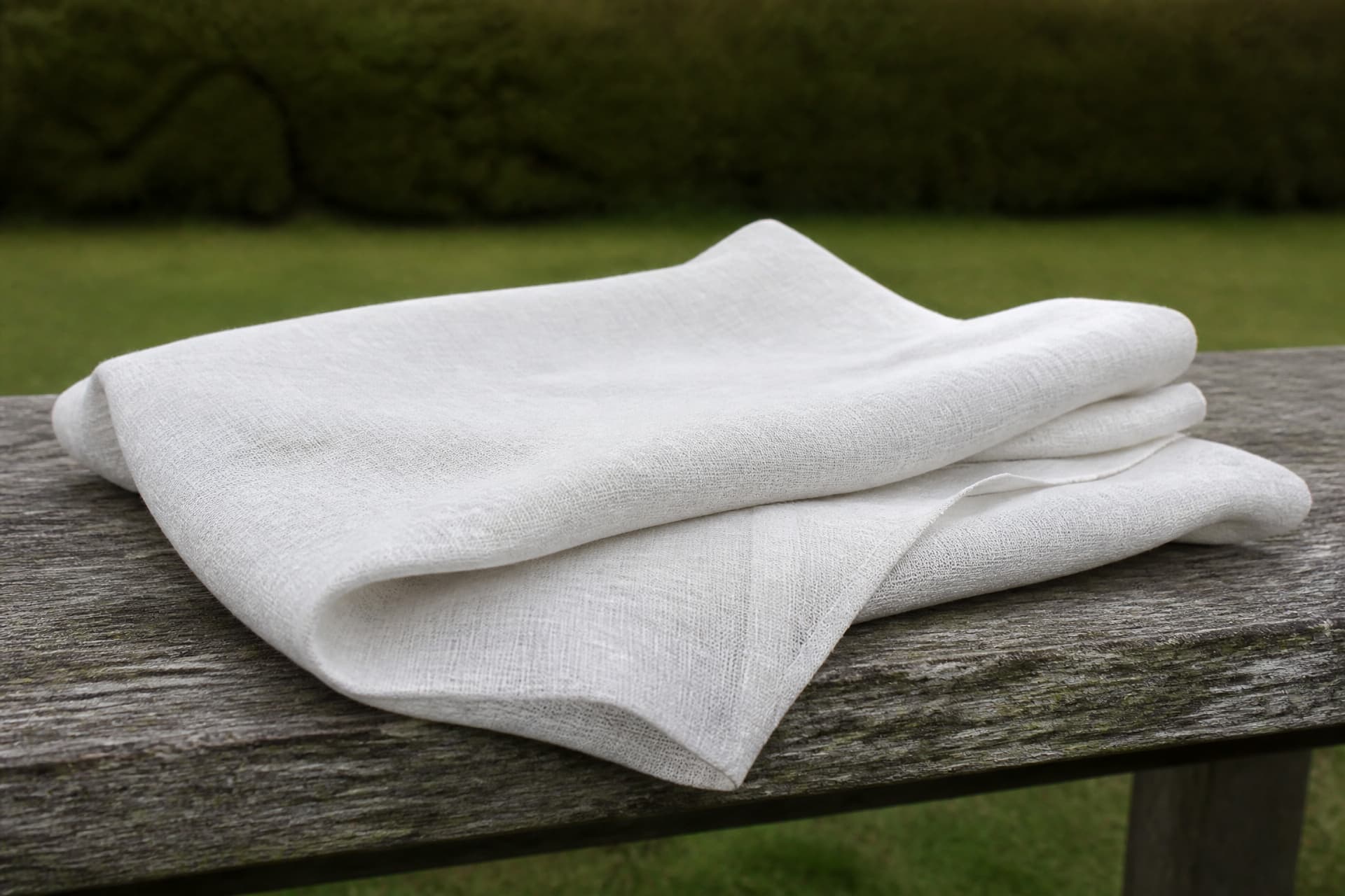 A single length of undyed white linen loosely folded on a weathered wooden bench outdoors, with out-of-focus grass behind it