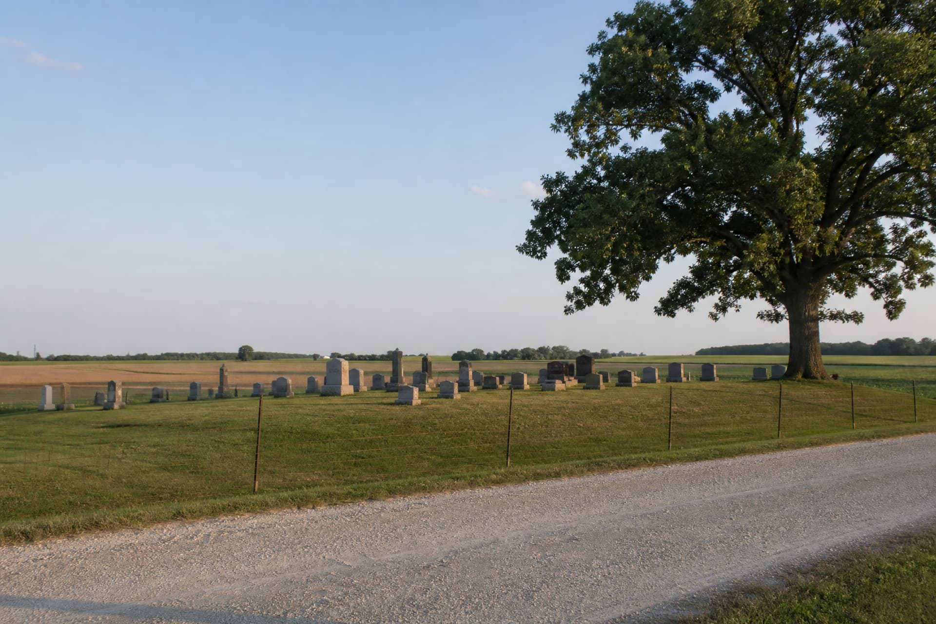A small rural cemetery on a low rise beside a gravel road in the American Midwest, a few dozen modest upright stones among mown grass, a plain wire fence and a single oak, with open farmland running to the horizon under soft afternoon light