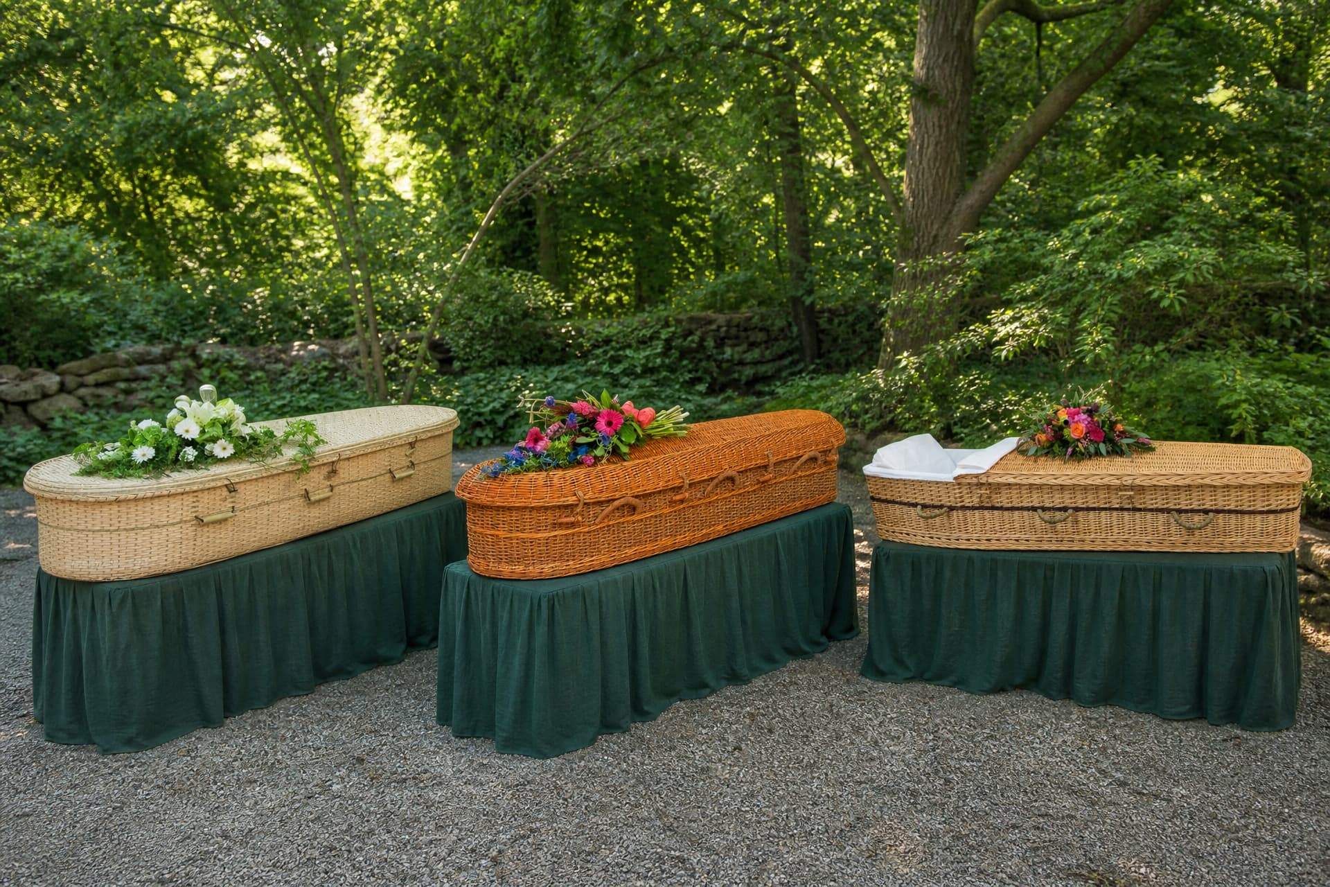 Three full-size woven caskets resting on draped green biers on a gravel path in a wooded setting, one pale bamboo, one honey-colored willow and one seagrass, each with a floral spray laid on the lid