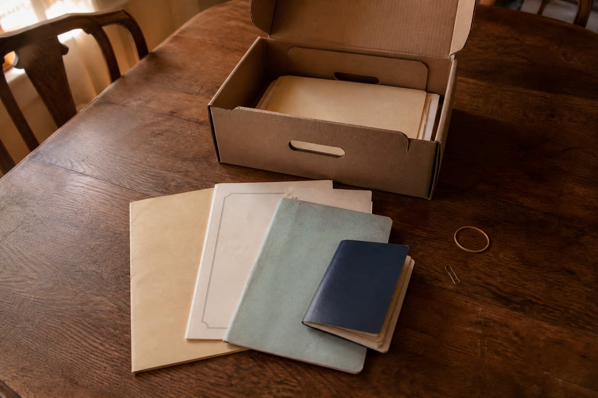An open cardboard document box on a dining table seen from above, old insurance policy folders and a savings passbook fanned out beside it in warm afternoon light