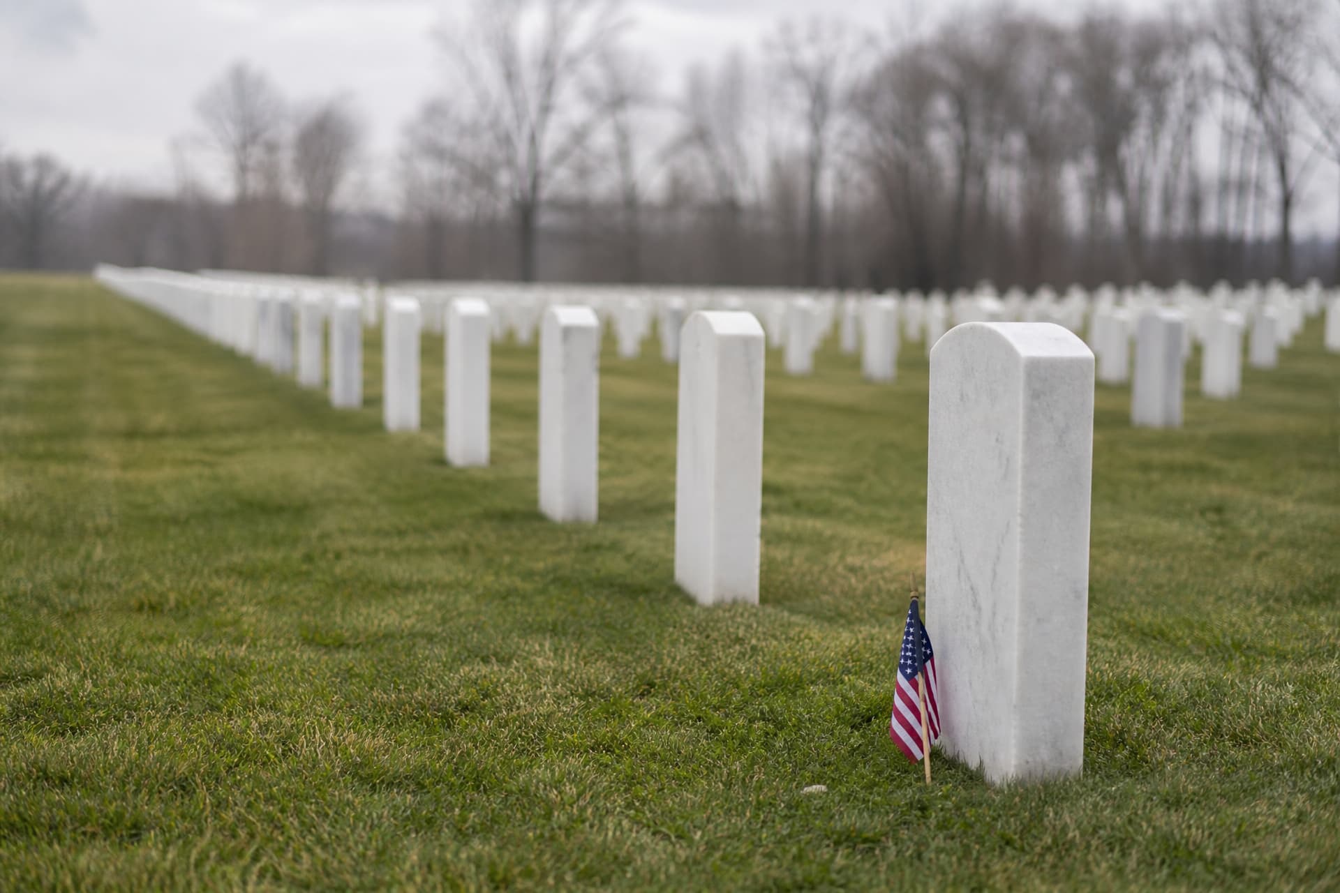 Rows of identical white upright headstones in a national veterans cemetery receding toward bare trees, mown grass and flat overcast morning light, a small flag at the base of the nearest stone
