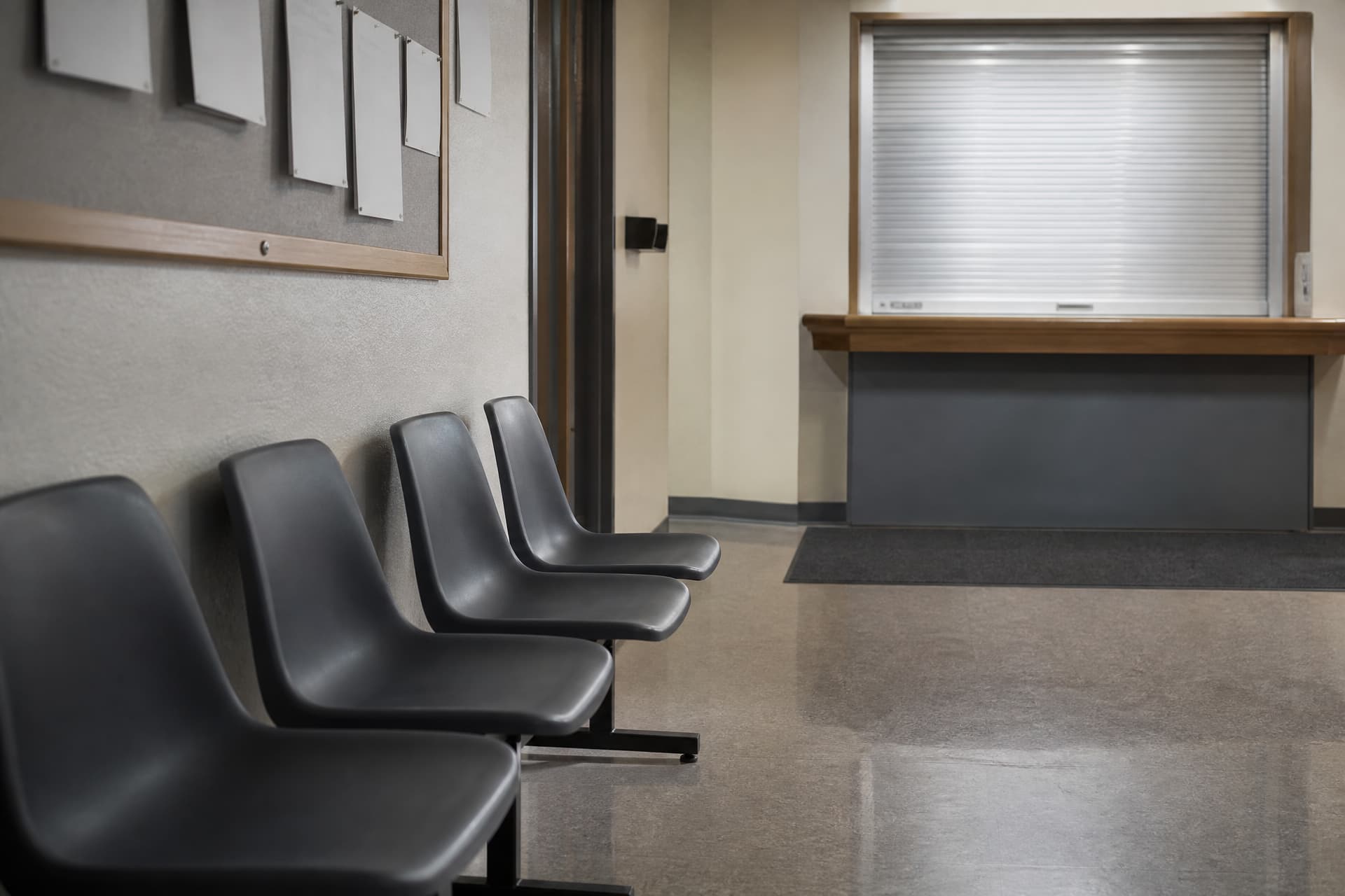 The waiting area of a small county government office, a row of moulded plastic chairs against a pale wall and a service counter with its window closed at the far end