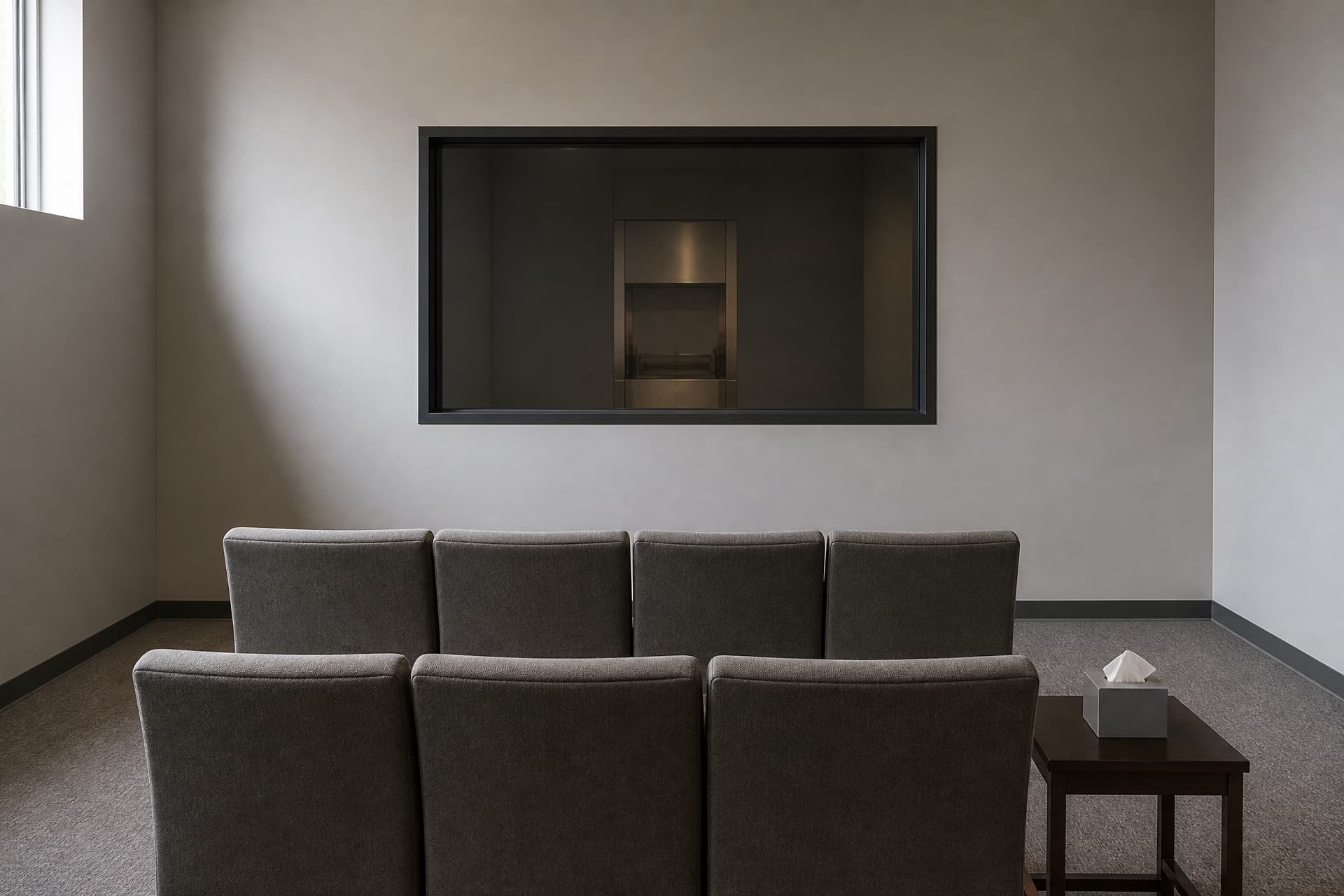 An empty crematory witness room in an American funeral home, with two rows of plain chairs facing a closed viewing window and a side table holding a box of tissues