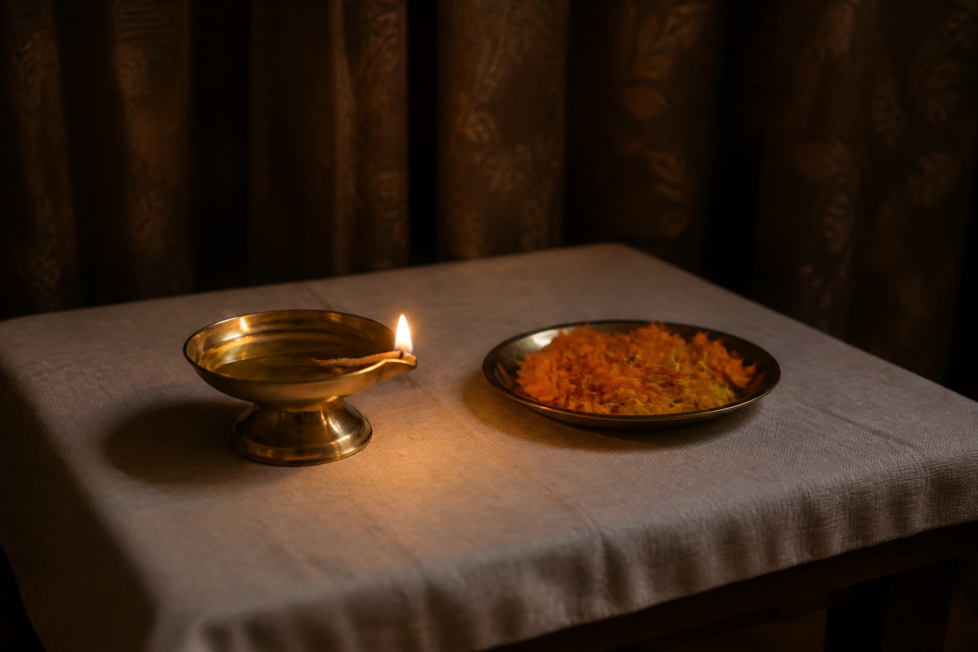 A small brass oil lamp burning on a low wooden table covered with white cloth, with a shallow brass dish of loose marigold petals beside it in a dim curtained room