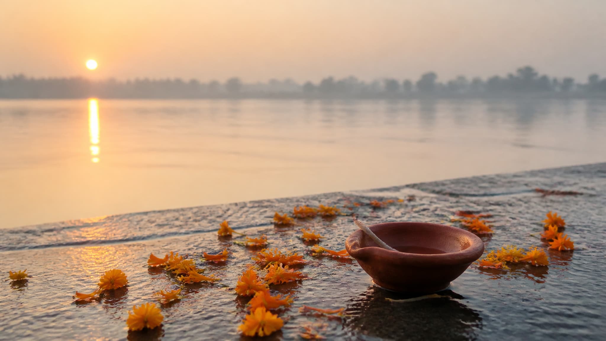 A wide river at first light seen from the bank, with loose marigold flowers and petals and a single unlit clay lamp resting on wet stone at the water's edge