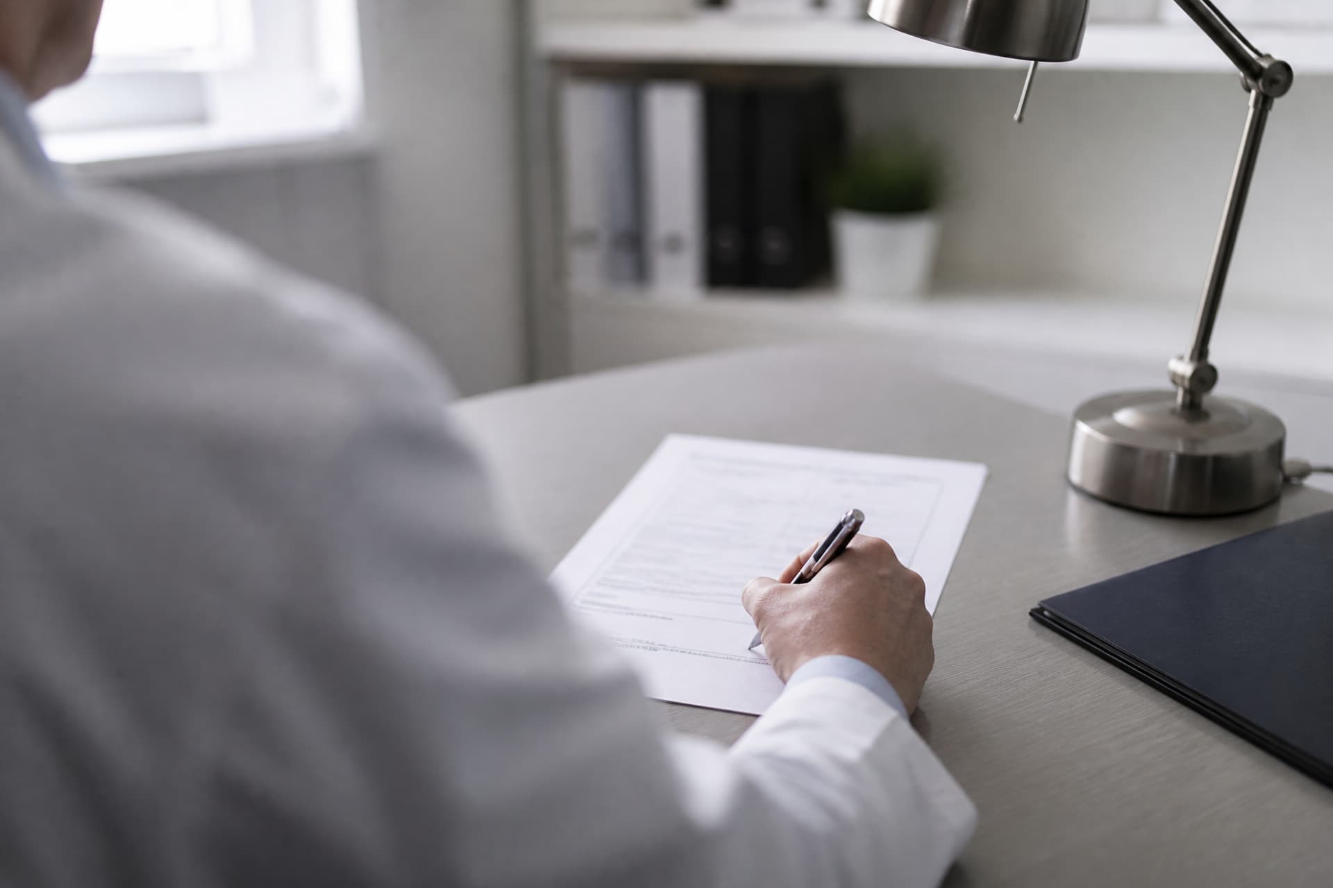 Over-the-shoulder view of a doctor in a white coat at a desk, signing a printed medical form with a pen, only the shoulder and hand in frame, a desk lamp and a closed folder beside the paper