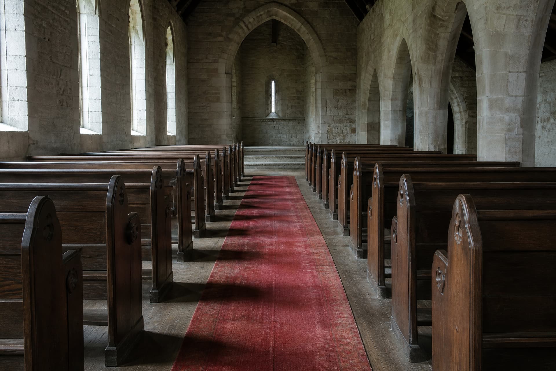 The center aisle of an empty church seen from the rear, wooden pews receding toward a stone chancel step, a narrow strip of worn red carpet down the middle in cool daylight