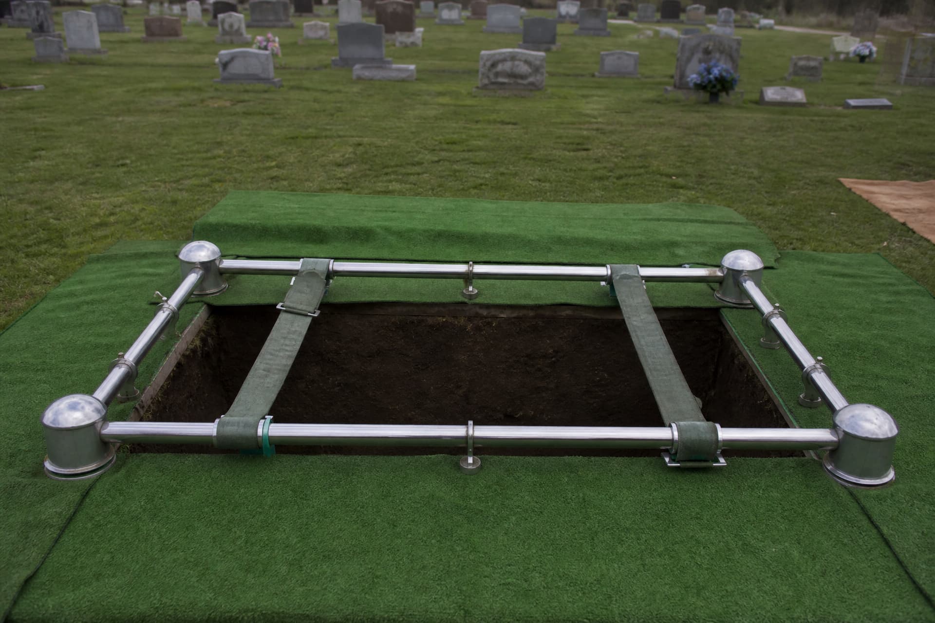 An open grave with a chrome lowering device set over it, webbing straps laid slack across the frame, artificial green turf covering the soil edges and headstones out of focus behind