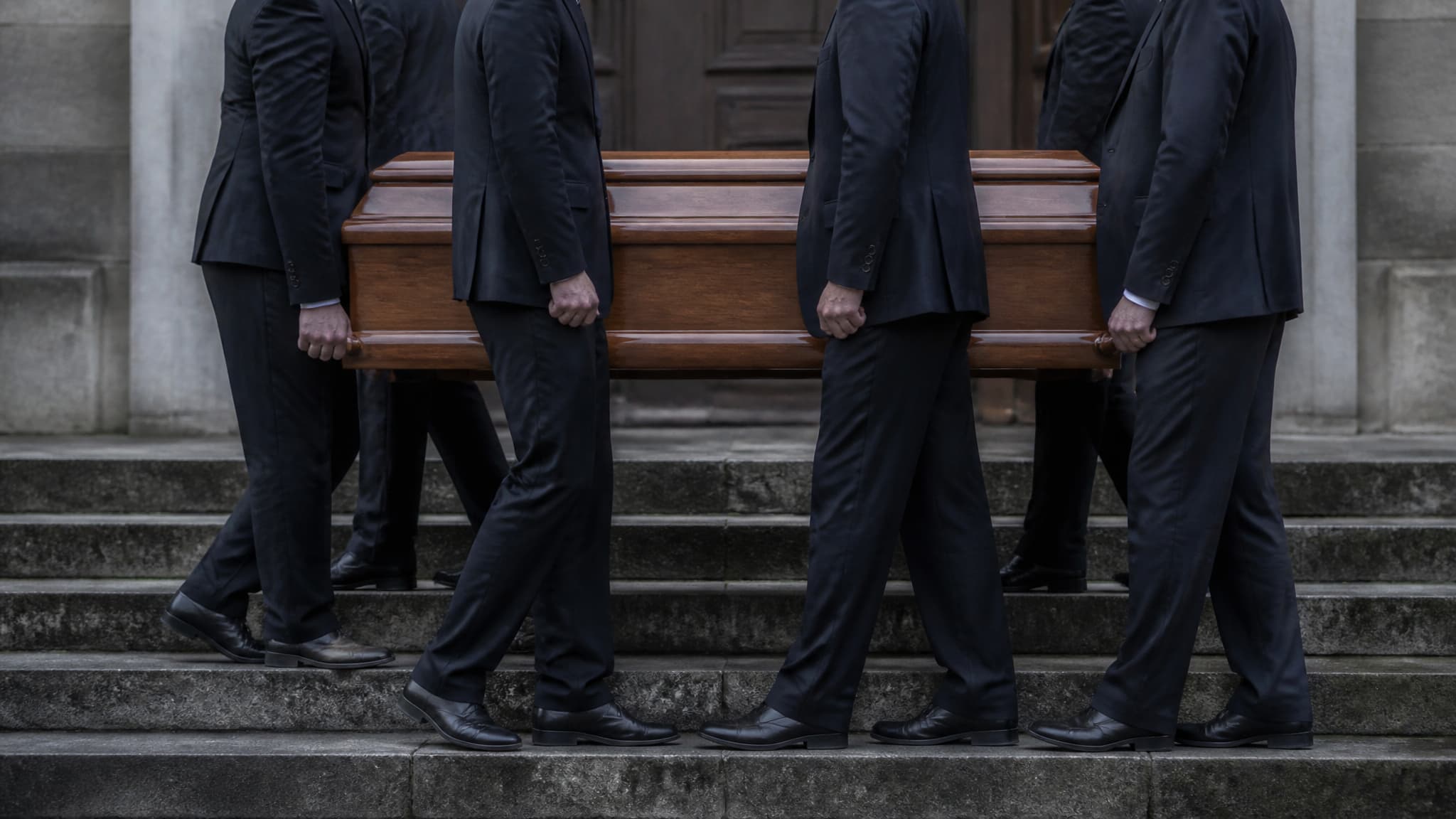 Six pallbearers in dark suits carrying a polished wooden casket down a short flight of stone church steps in overcast daylight, hands gripping the side rails, no faces visible above the shoulders
