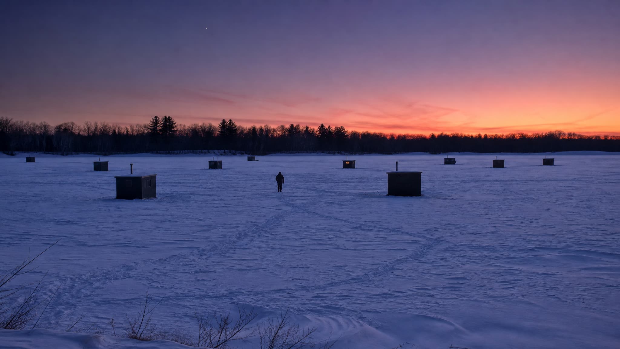 A frozen Minnesota lake at dusk, a dozen small dark ice-fishing houses scattered across the wind-scoured snow, packed tracks curving between them, one figure walking alone, and a dark treeline under an orange and violet sky
