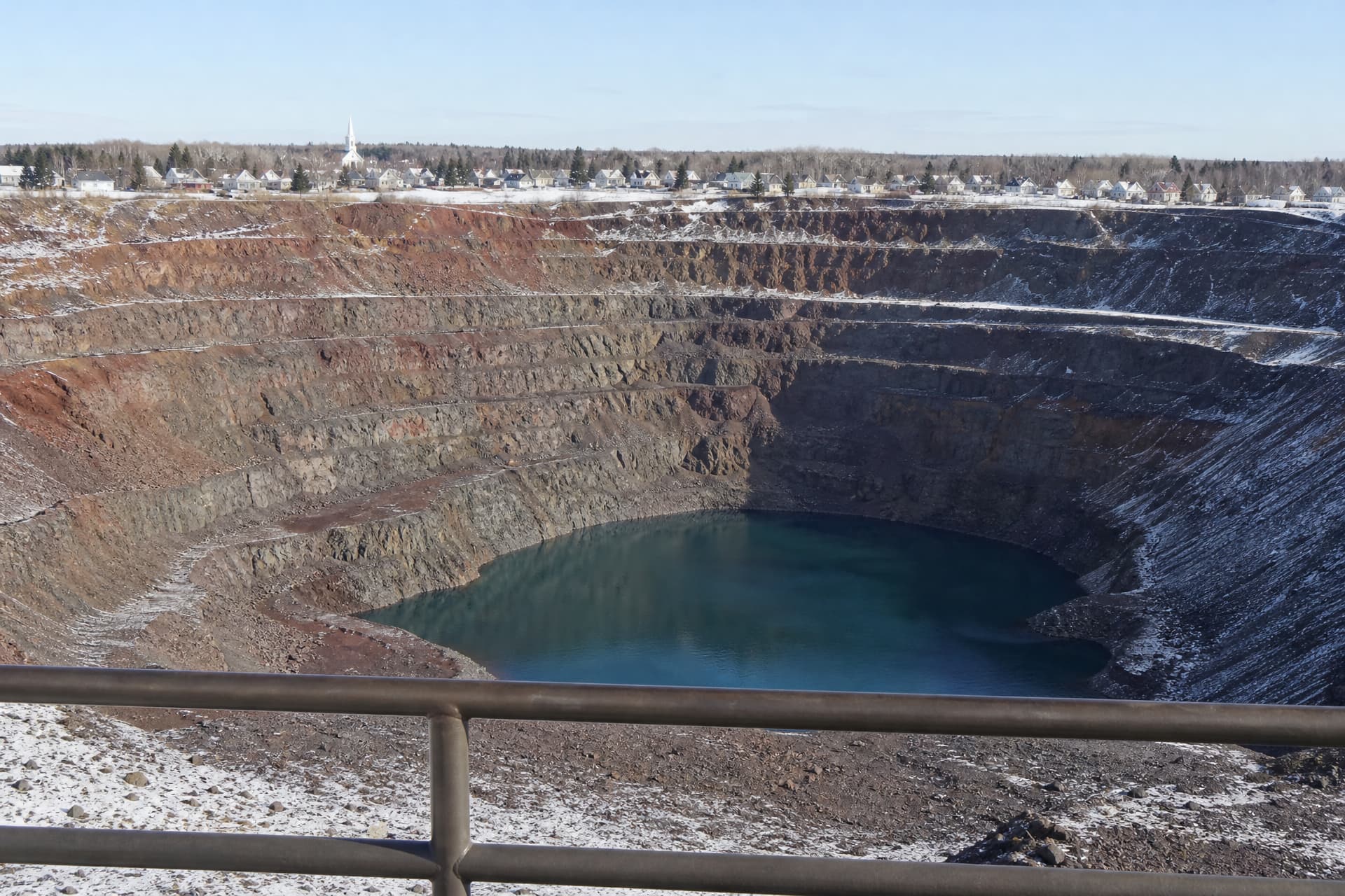 A terraced open-pit iron mine on Minnesota's Iron Range seen from its rim behind a steel guardrail, rust-red rock benches stepping down to blue-green water, and a small town of white houses and a church spire along the far rim