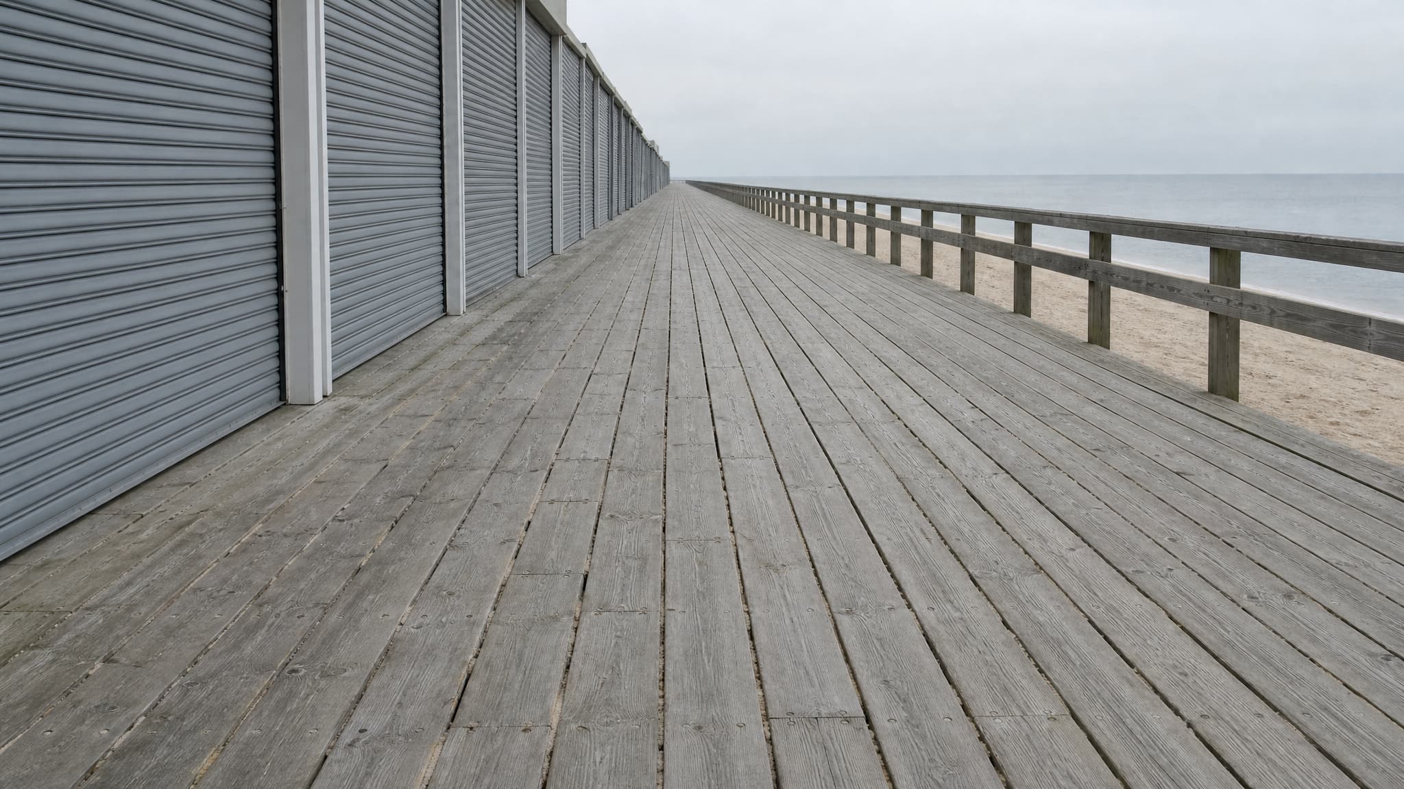 An empty Jersey Shore boardwalk in the off season, weathered silver planking running away from the camera past a long run of closed metal shutters, with a wooden rail, pale beach and flat gray sea to the right