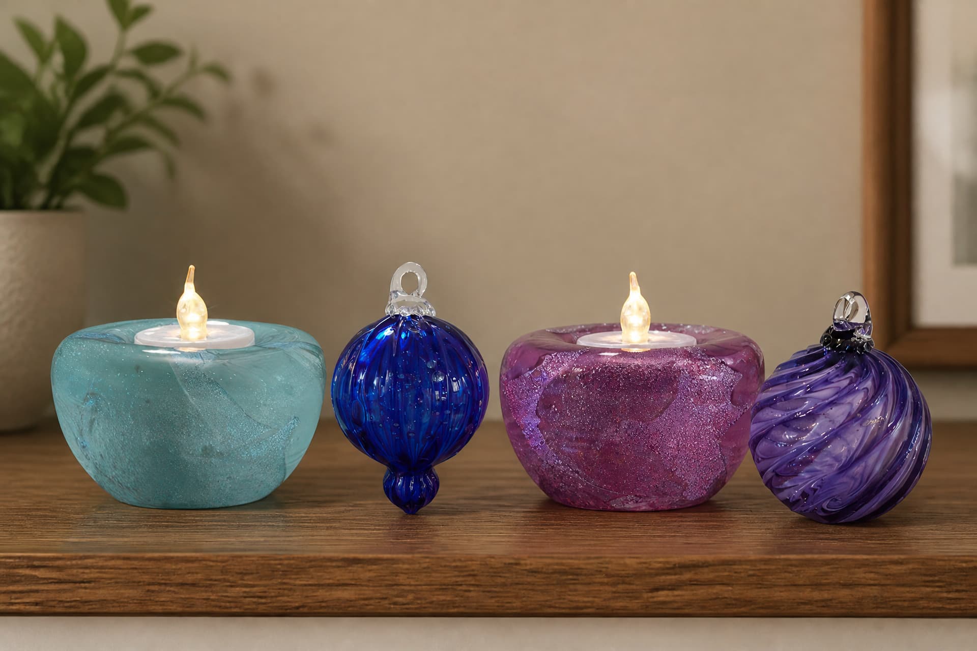 Two lit glass tealight holders in aqua and pink standing on a wooden shelf, with a ribbed cobalt blue glass bauble and a purple swirled glass ornament beside them