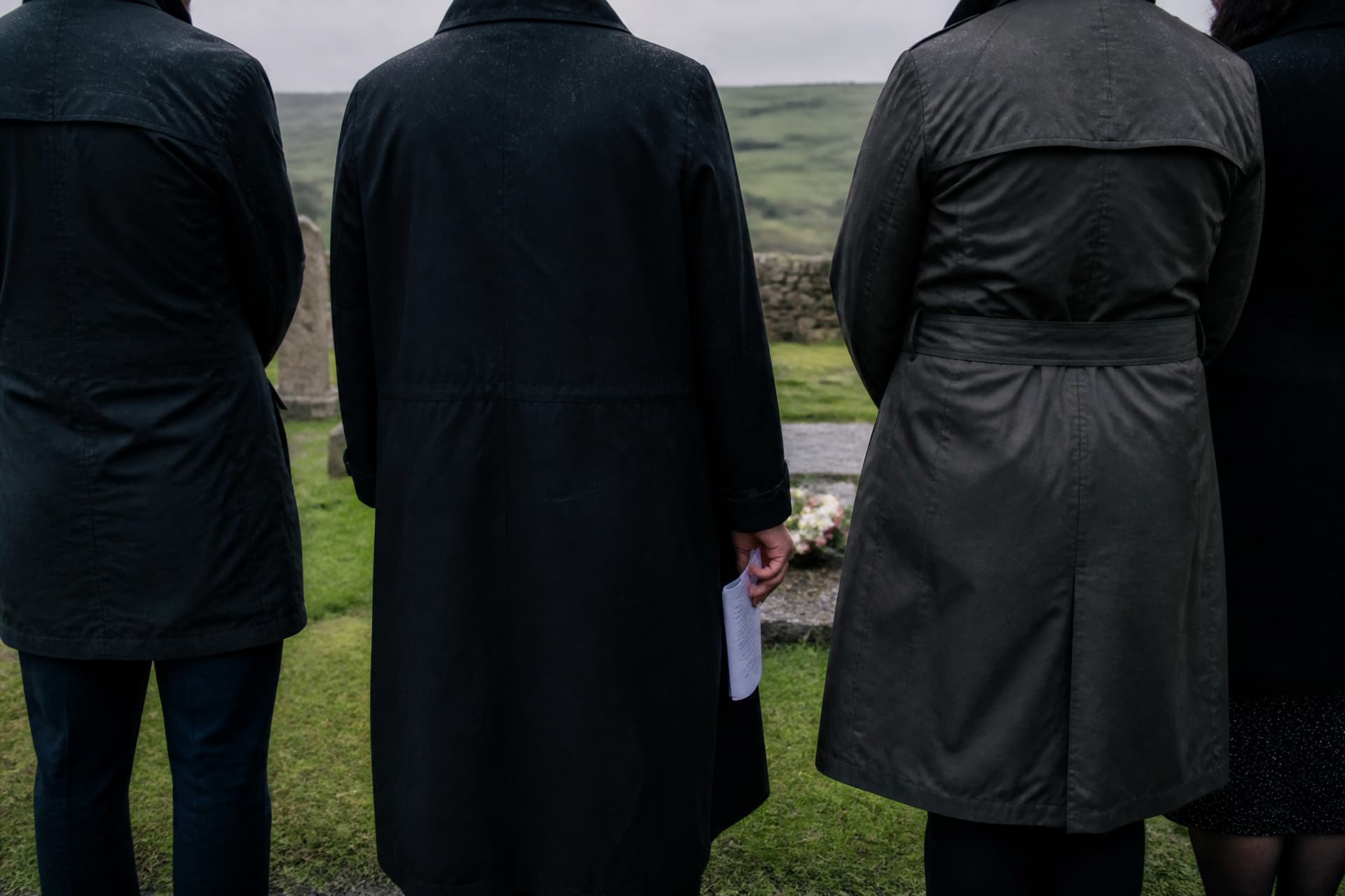 Four mourners in dark coats seen from behind at a graveside in an Irish churchyard, one holding a folded order of service at their side, a low stone wall and green hills beyond under drizzle
