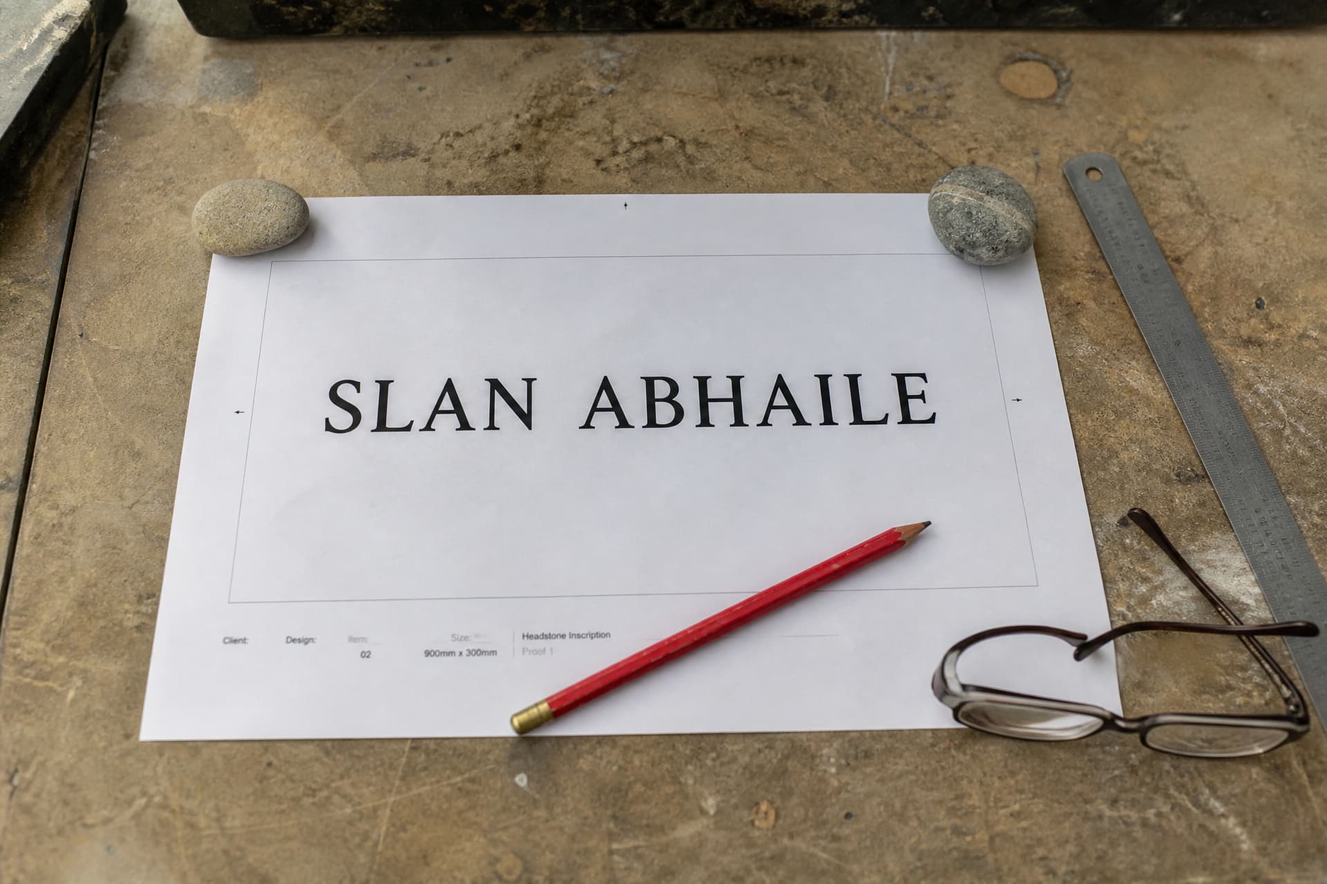 A full-size paper proof of a headstone inscription weighted down on a stone workshop bench, the lettering reading SLAN ABHAILE with a red pencil, a steel rule and reading glasses beside it