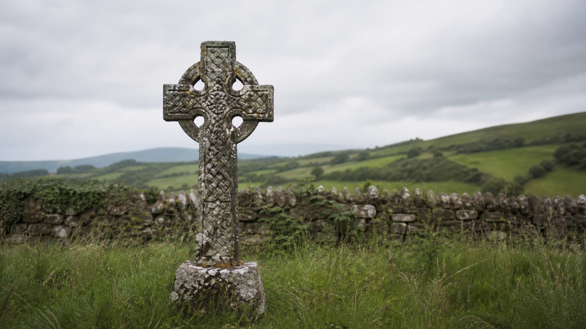 A weathered granite Celtic cross standing in long grass in an Irish country churchyard under a soft gray sky, lichen on the stone and low green hills beyond the wall