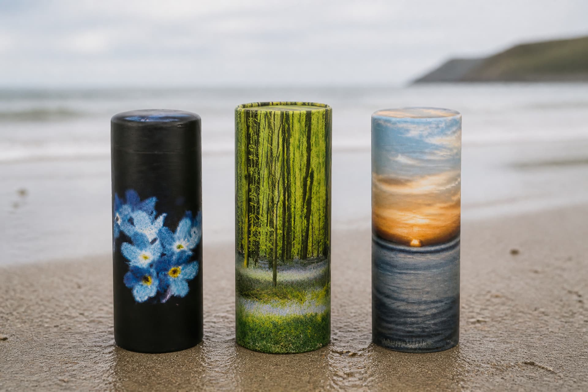Three cylindrical scattering urns standing on firm damp sand at the water's edge, printed with a forget-me-not design, a woodland scene and an ocean sunset, with breaking surf and a headland soft behind them in overcast light