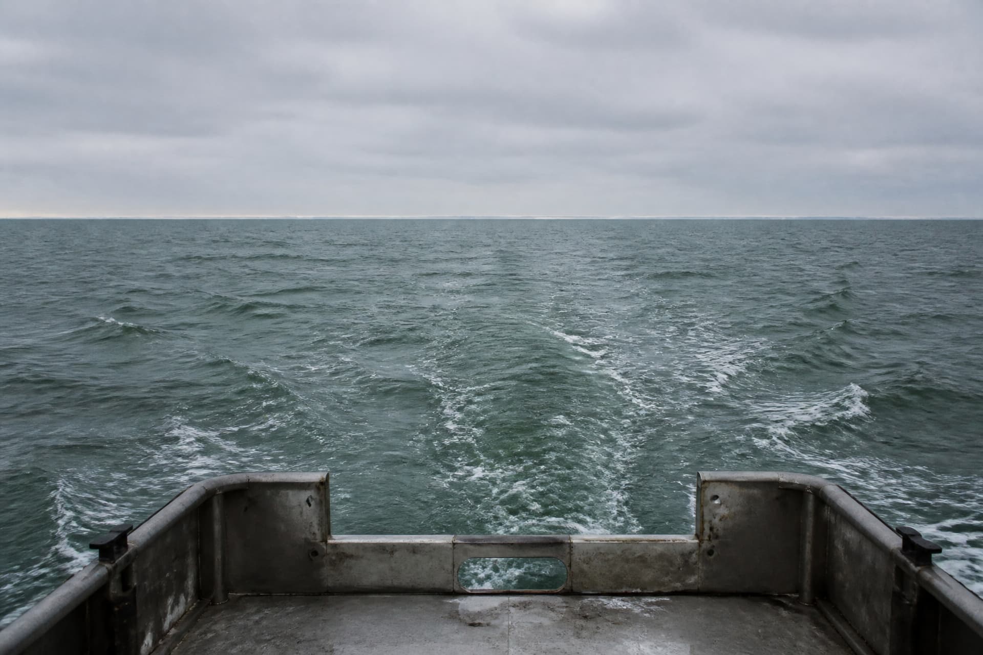 A view from the stern of a small boat looking back toward a distant low coastline reduced to a thin pale line on the horizon under flat gray sky, open green-gray water filling most of the frame with a faint wake