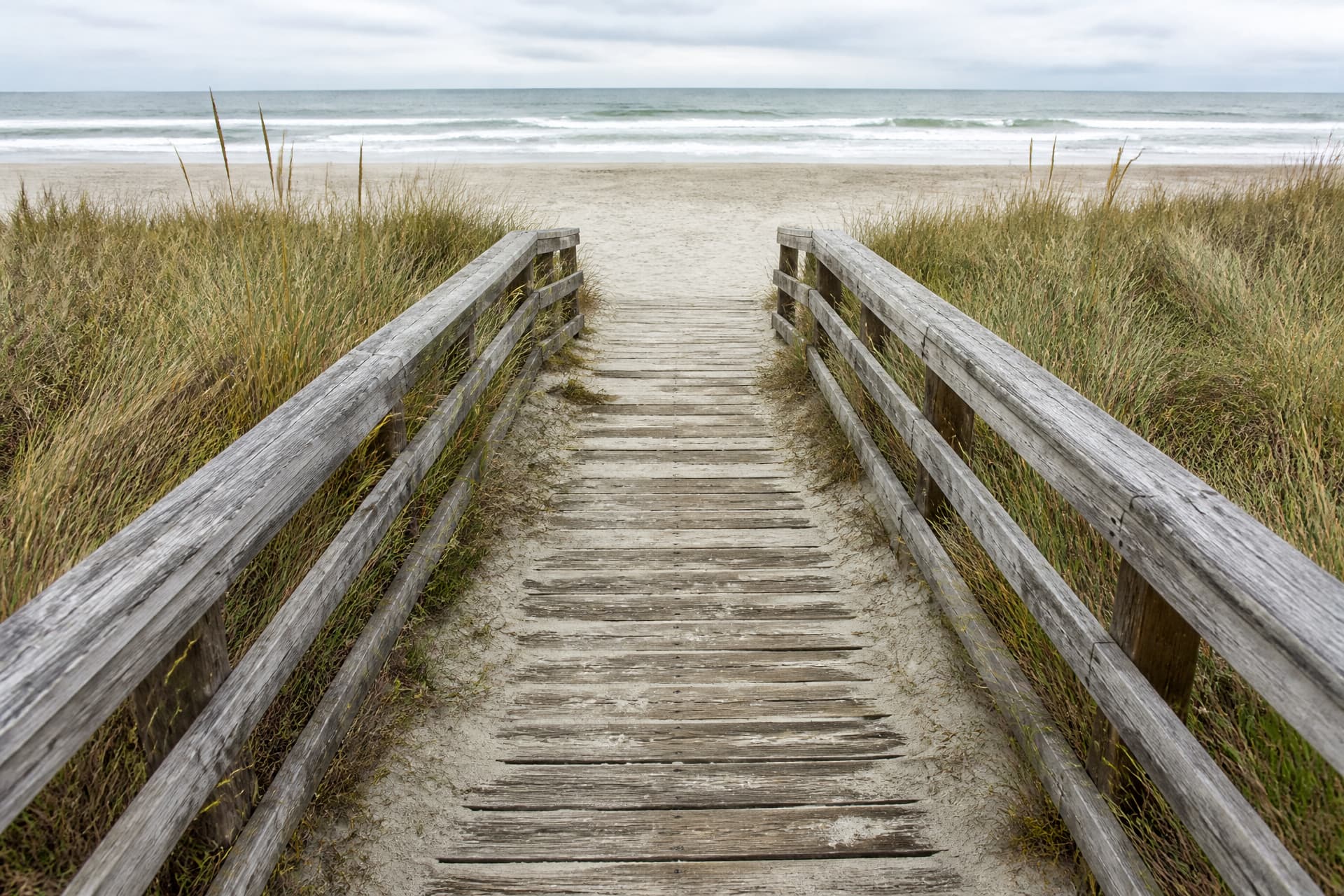 A weathered wooden beach access boardwalk with slatted timber rails crossing low dune grass toward open sand and breaking surf, photographed down its length in flat overcast daylight