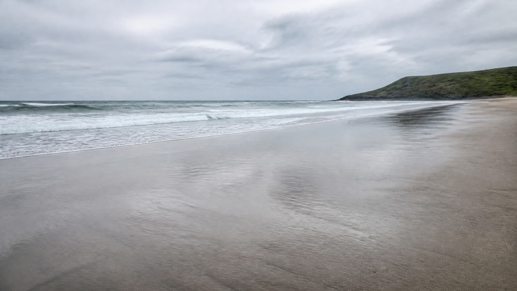 A wide empty sand beach at low tide in flat overcast morning light, wet sand in the foreground reflecting a pale sky, gentle surf a short distance out and a low headland at the far right of the frame