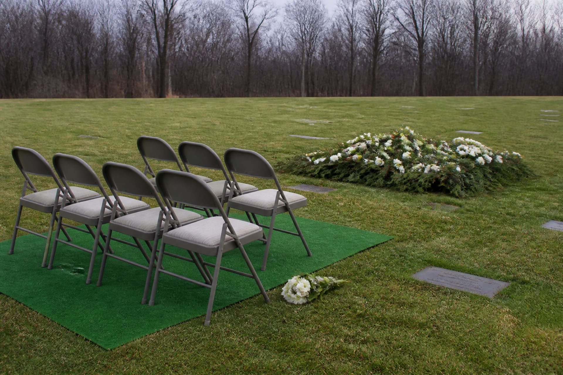 Empty folding chairs on green matting beside a flower-covered grave in an ordinary cemetery under overcast light