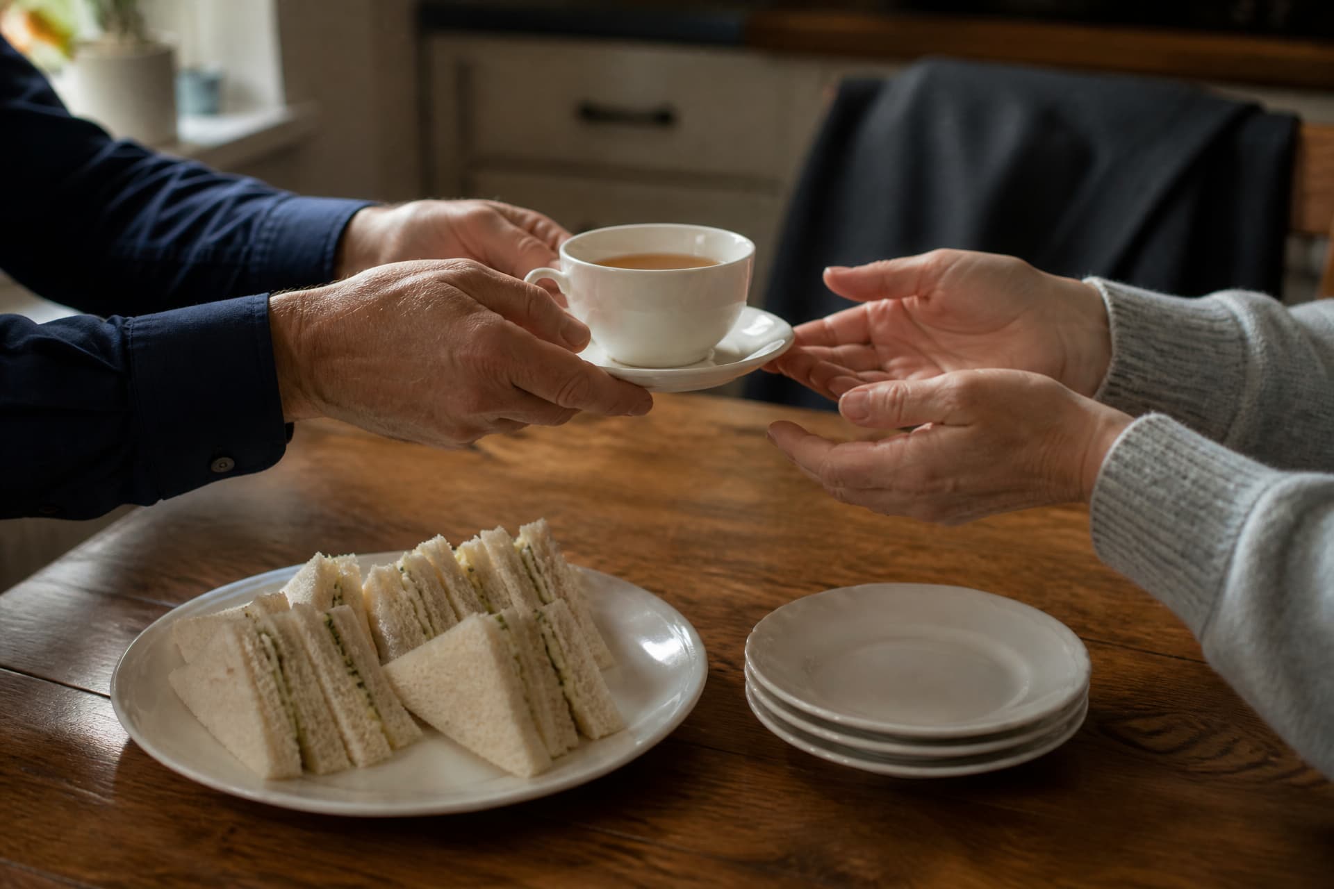 One pair of hands passing a cup of tea across a kitchen table to another pair of hands, with a plate of simple food between them and a folded dark jacket over a chair back