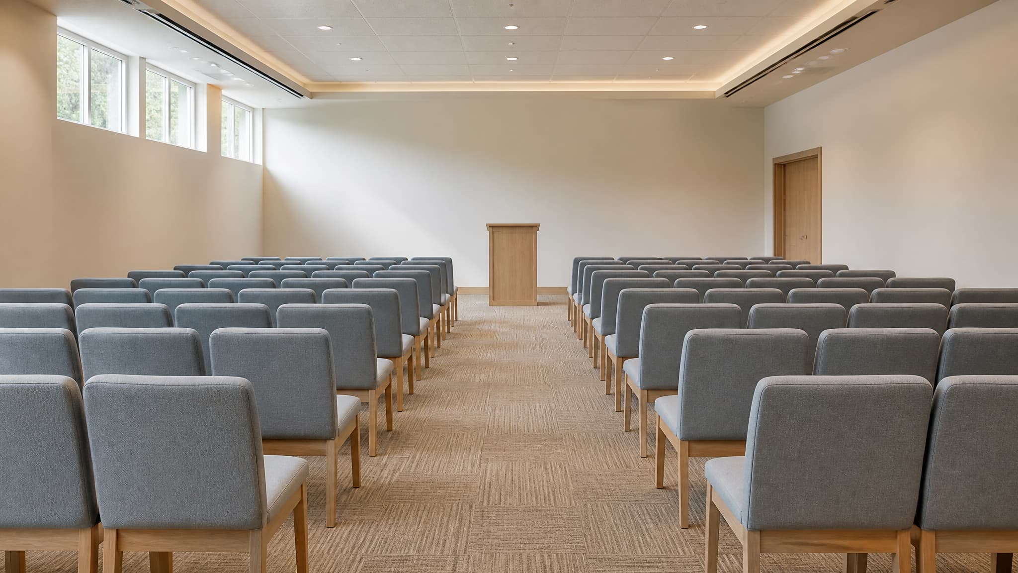 Rows of plain upholstered chairs facing a low wooden lectern in a simple meeting hall lit by high windows
