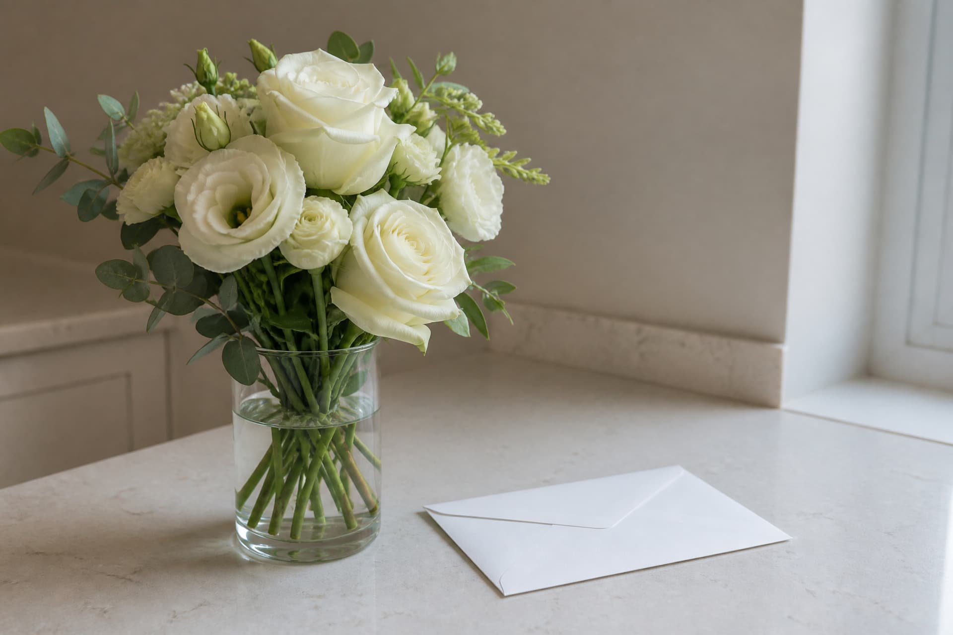 A small hand-tied arrangement of white and cream flowers in a plain glass vase on a kitchen counter beside a sealed envelope