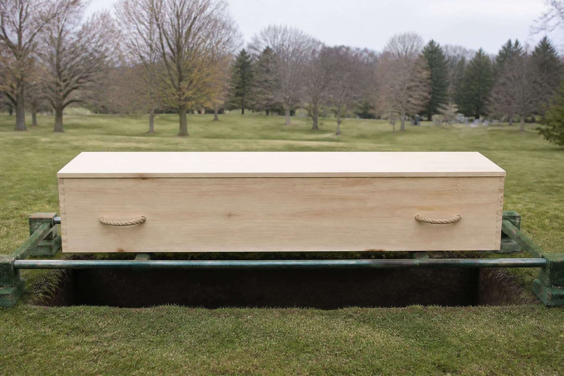 A plain unfinished pine box with a flat panel lid, visible corner joinery and rope side handles, resting on lowering rails over an open grave in mown grass under overcast daylight