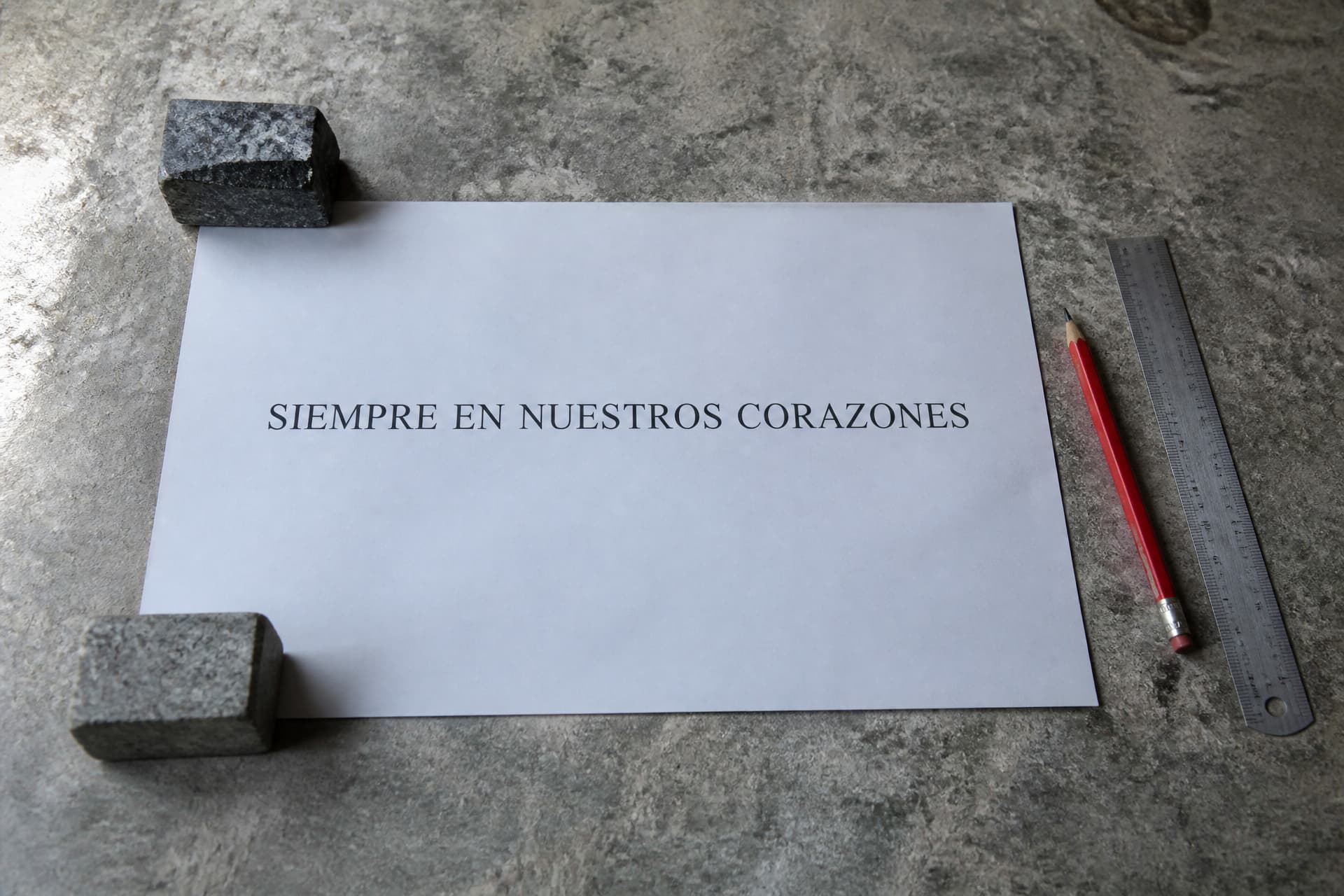 Una prueba impresa a tamaño real sobre el banco de un taller de piedra, sujeta con dos recortes de granito, con una sola línea en mayúsculas que dice SIEMPRE EN NUESTROS CORAZONES, y a un lado un lápiz rojo y una regla metálica