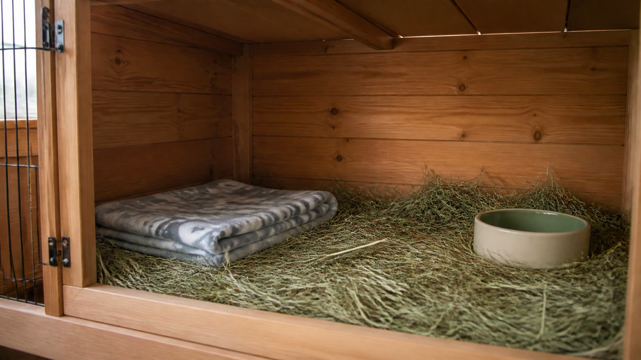 An empty hay-lined hutch corner in soft daylight with a small folded blanket inside