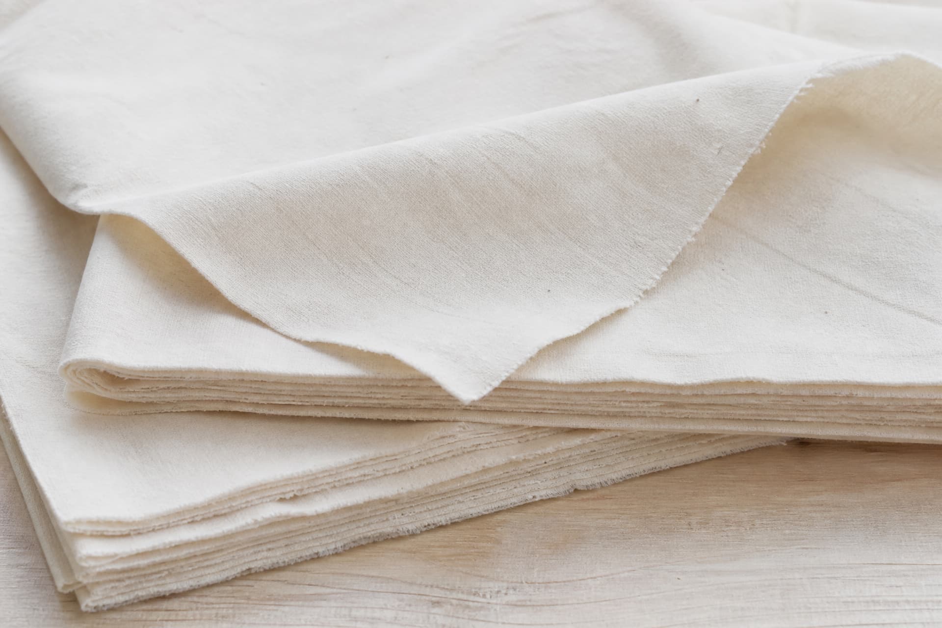 A close view of plain white unbleached cotton cloth folded in a stack, one raw-edged sheet falling open to show the weave