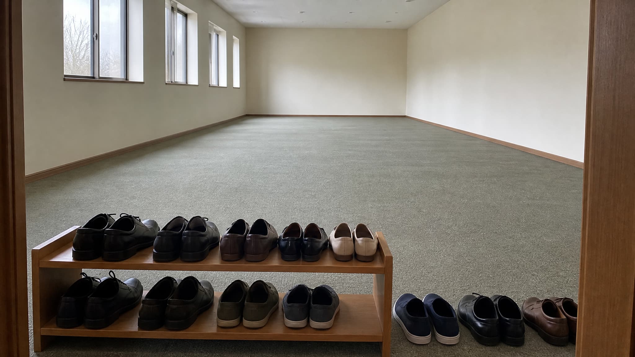 A low wooden shoe rack holding rows of ordinary shoes at the entrance to a plain, empty carpeted prayer hall in soft daylight