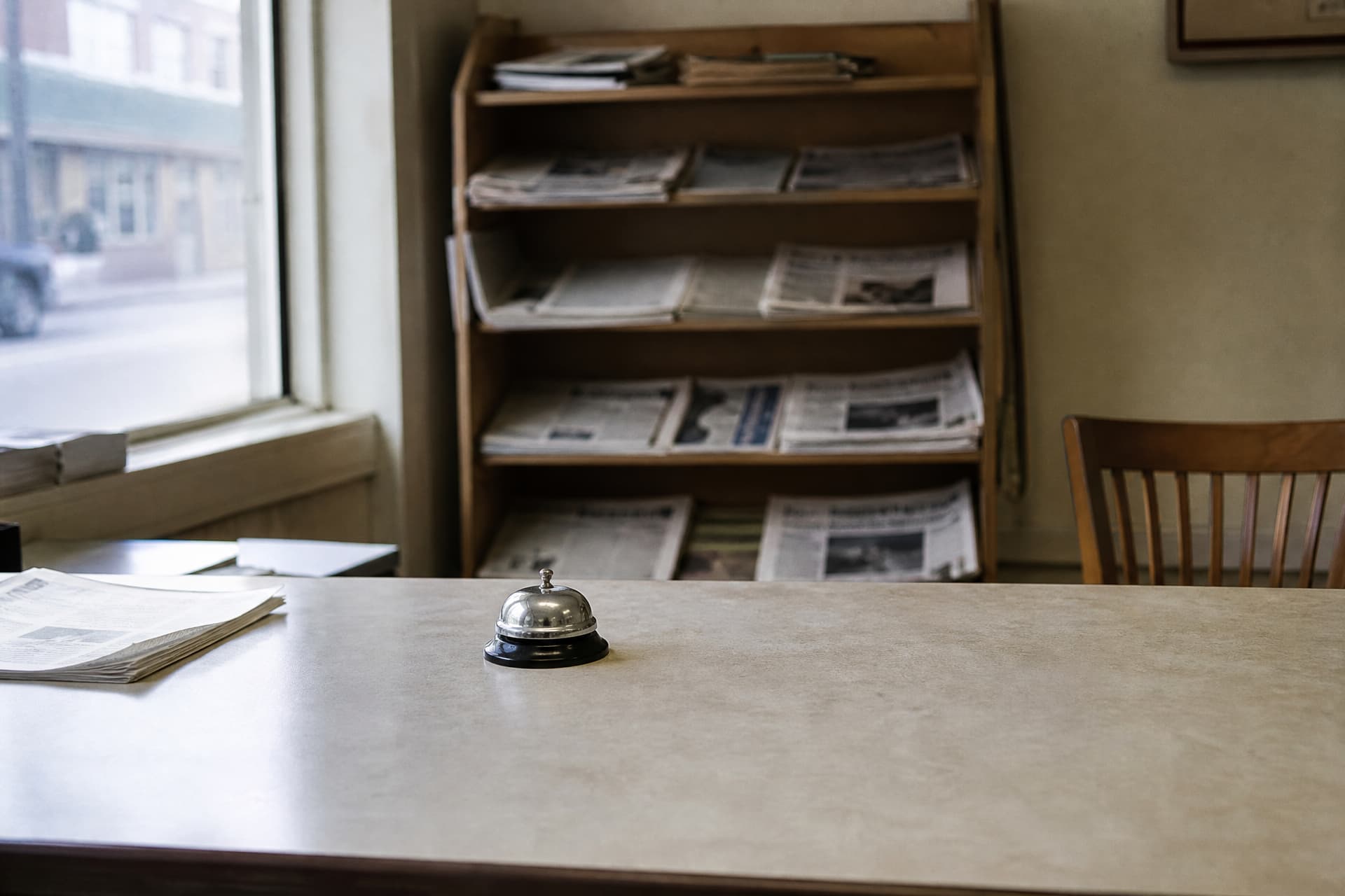 The public counter of a small local newspaper office, a bell on the counter, a rack of back issues behind and a wooden chair to one side in flat daylight