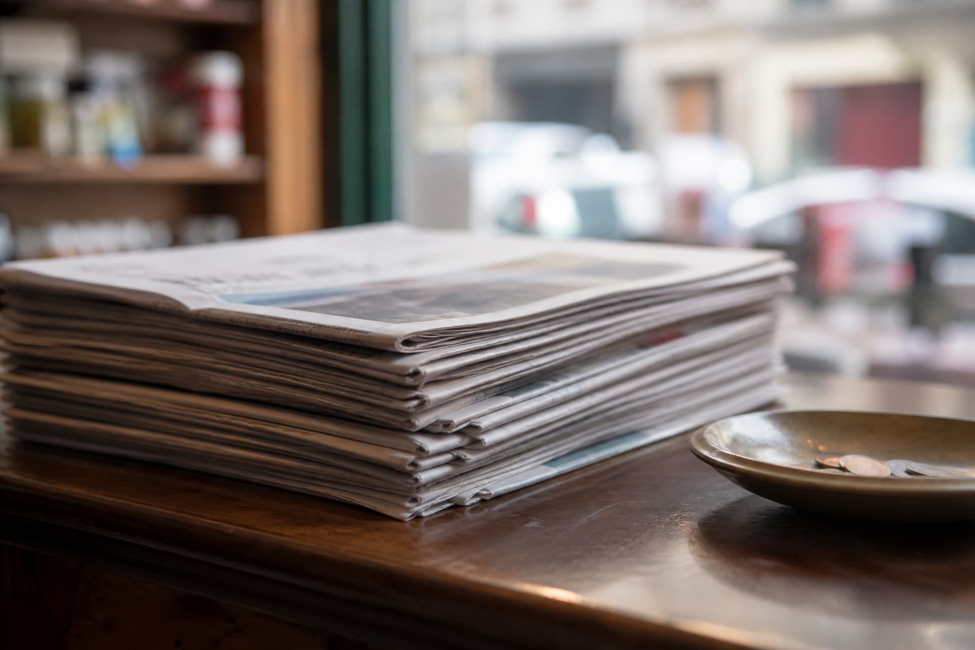 A stack of folded local newspapers on a shop counter seen from the side, a coin dish beside them, early morning light through a shopfront window