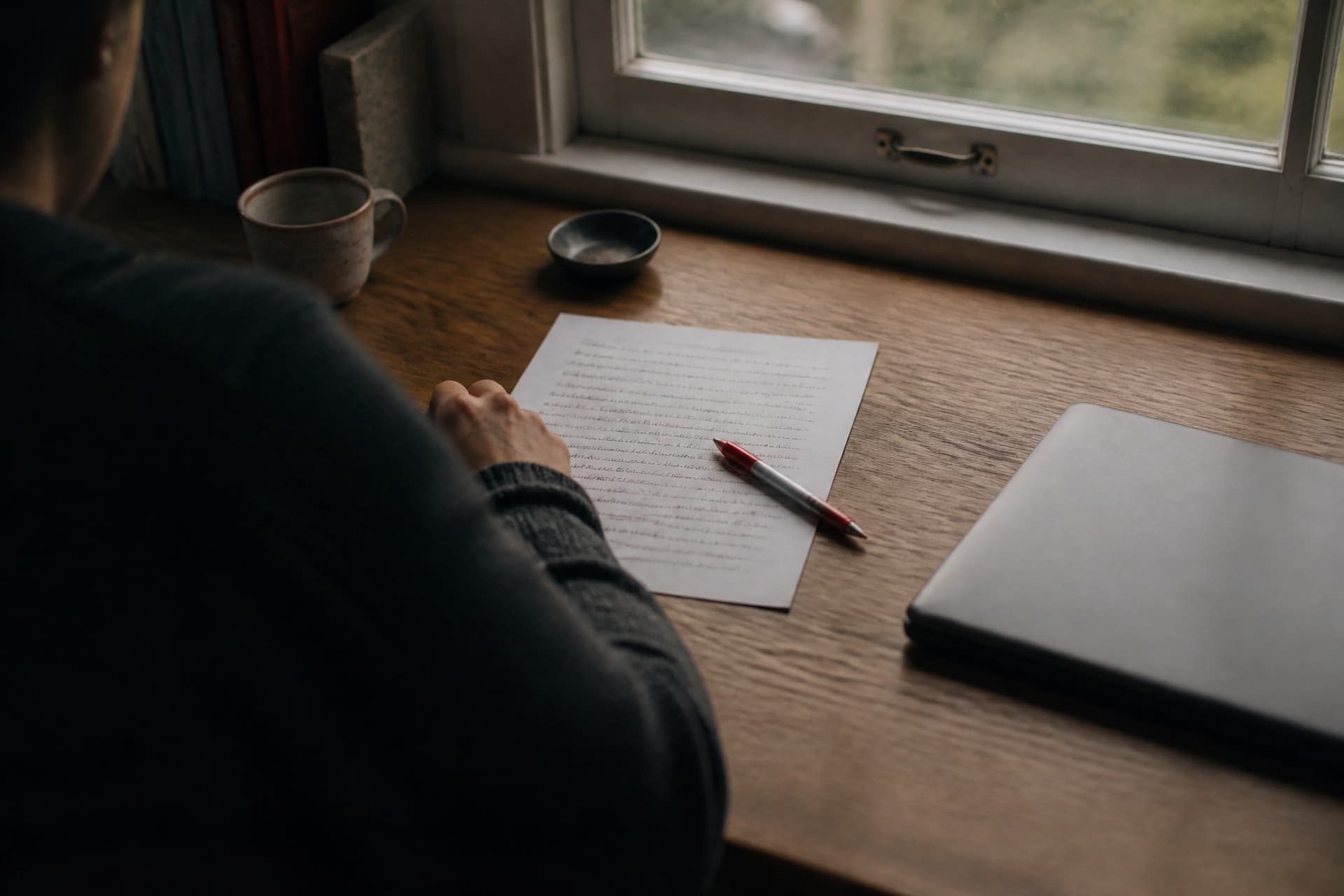 A writing desk by a window seen over the shoulder, a sheet of paper part-covered in handwriting with several lines crossed out in red pen, the pen resting on the page
