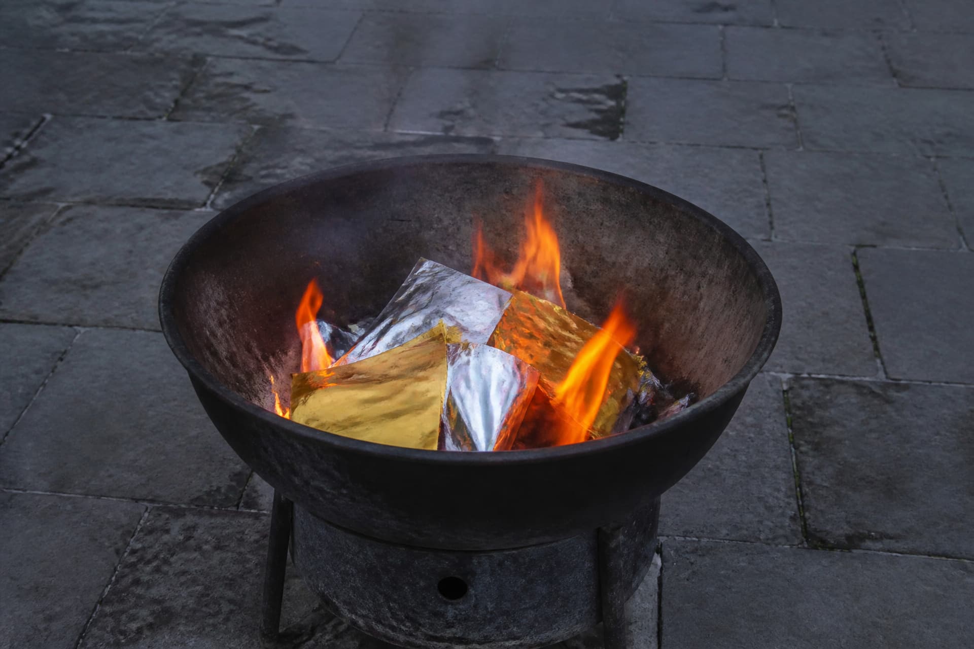 Folded sheets of plain gold and silver paper burning in a blackened metal brazier on a stone courtyard floor, soft smoke rising, warm firelight against cool gray stone, seen from standing height looking down
