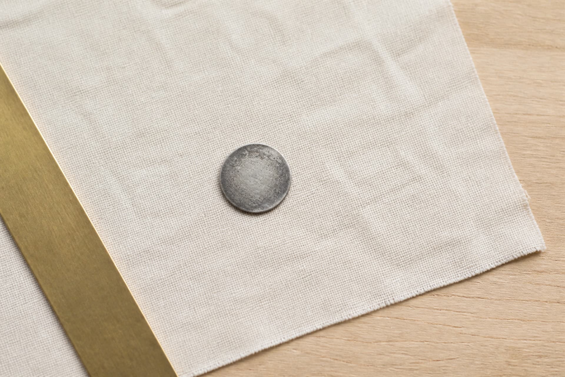 One small tarnished silver coin resting on plain cream linen cloth on a pale wood surface, photographed from close overhead in soft even daylight with a plain unmarked brass edge just visible at the side of the frame