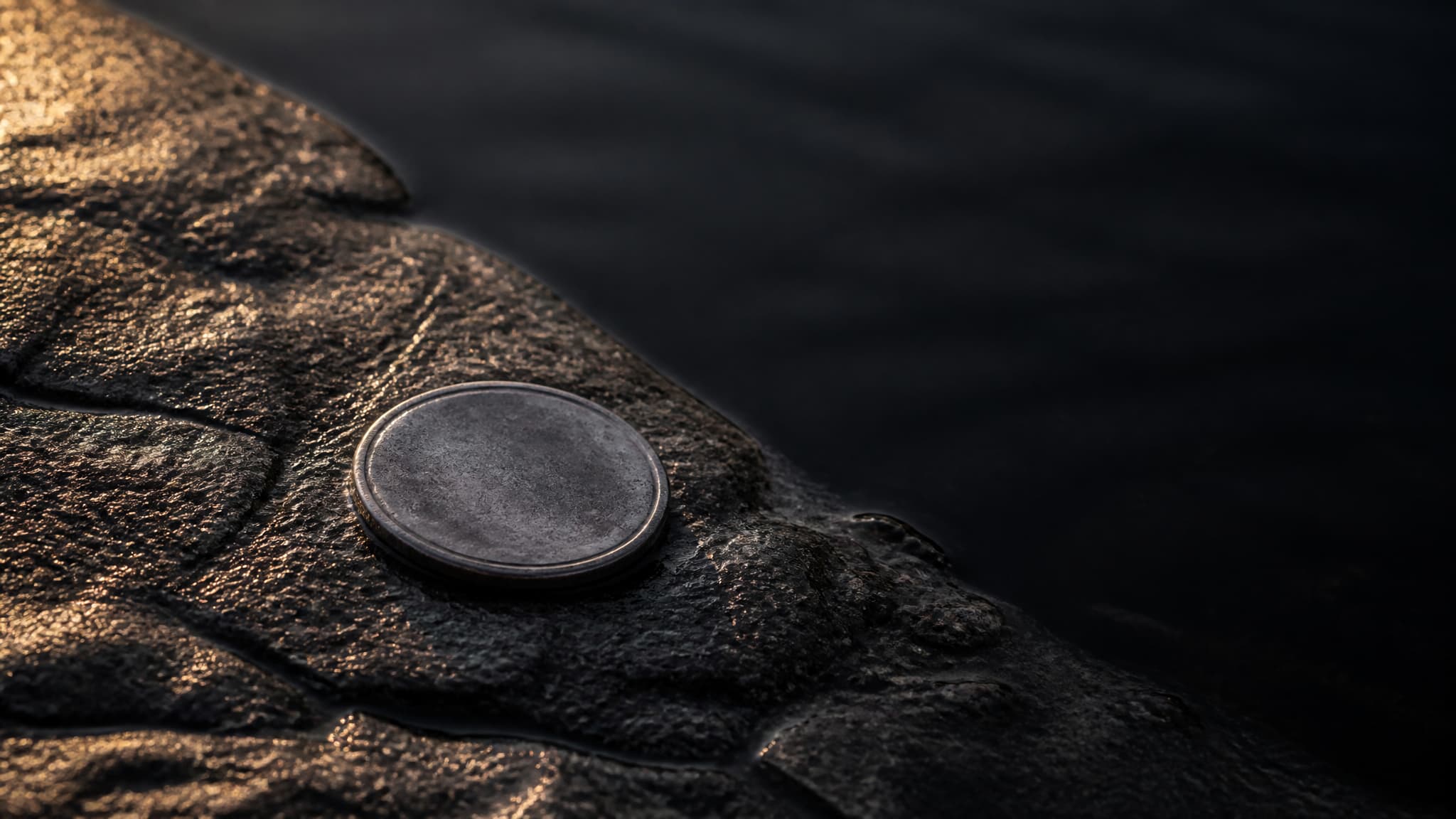 A single small worn silver coin lying on wet dark stone at the edge of still black water, lit by low warm light from one side with deep shadow beyond