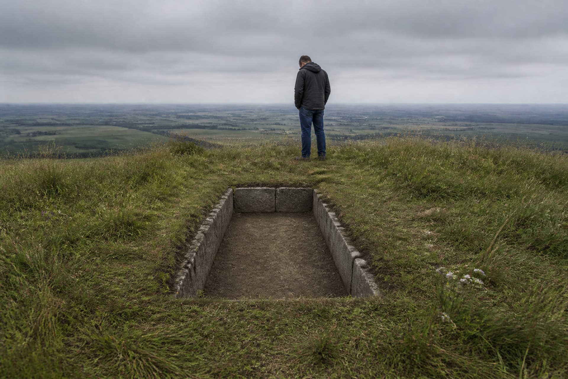 A shallow empty stone-lined grave cut into turf on an open European hilltop, its stones set deliberately in a rectangle, with a man in a dark jacket standing beyond it looking down into the grave under flat gray daylight
