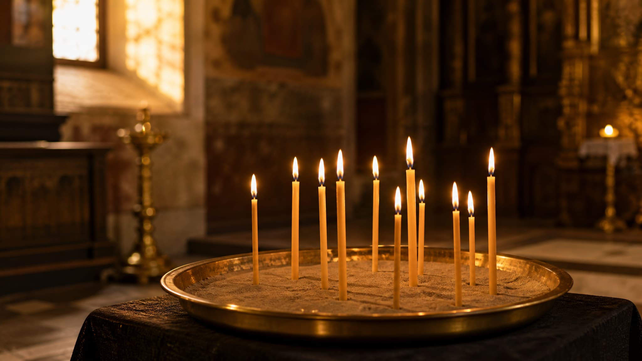 Slender lit beeswax candles standing in a tray of sand on a low memorial table inside an Orthodox church, late afternoon light through a high window